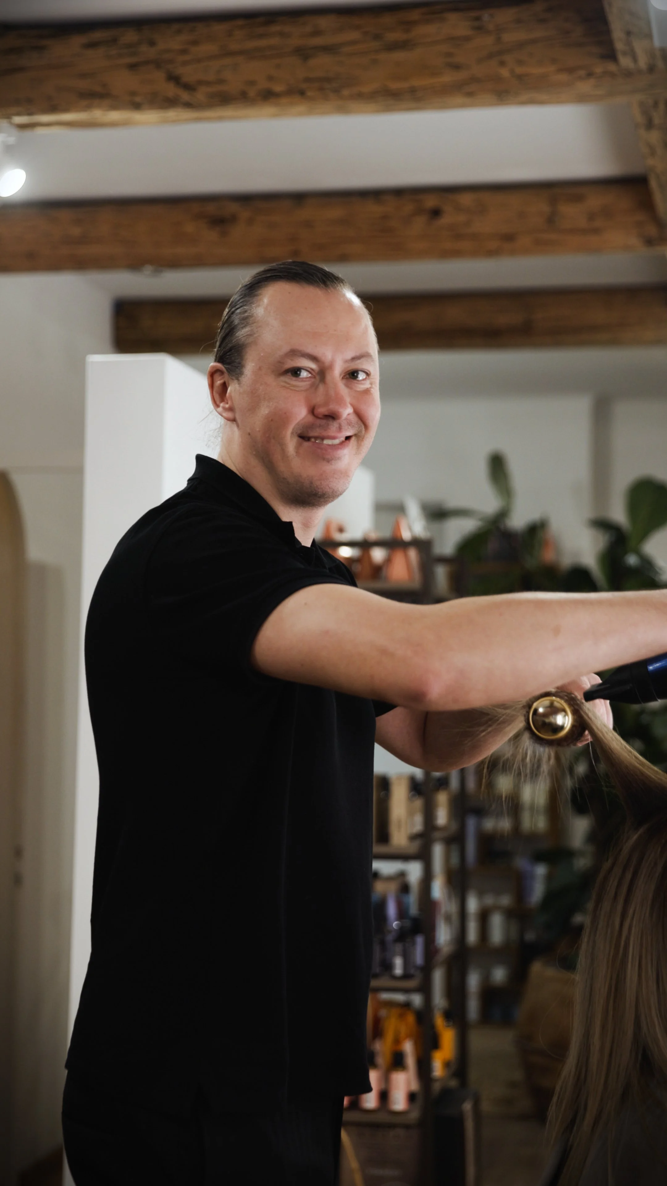 A man with shoulder-length brown hair and fair skin standing with arms crossed in front of large green leaves, wearing a black short-sleeve shirt and a black digital watch.