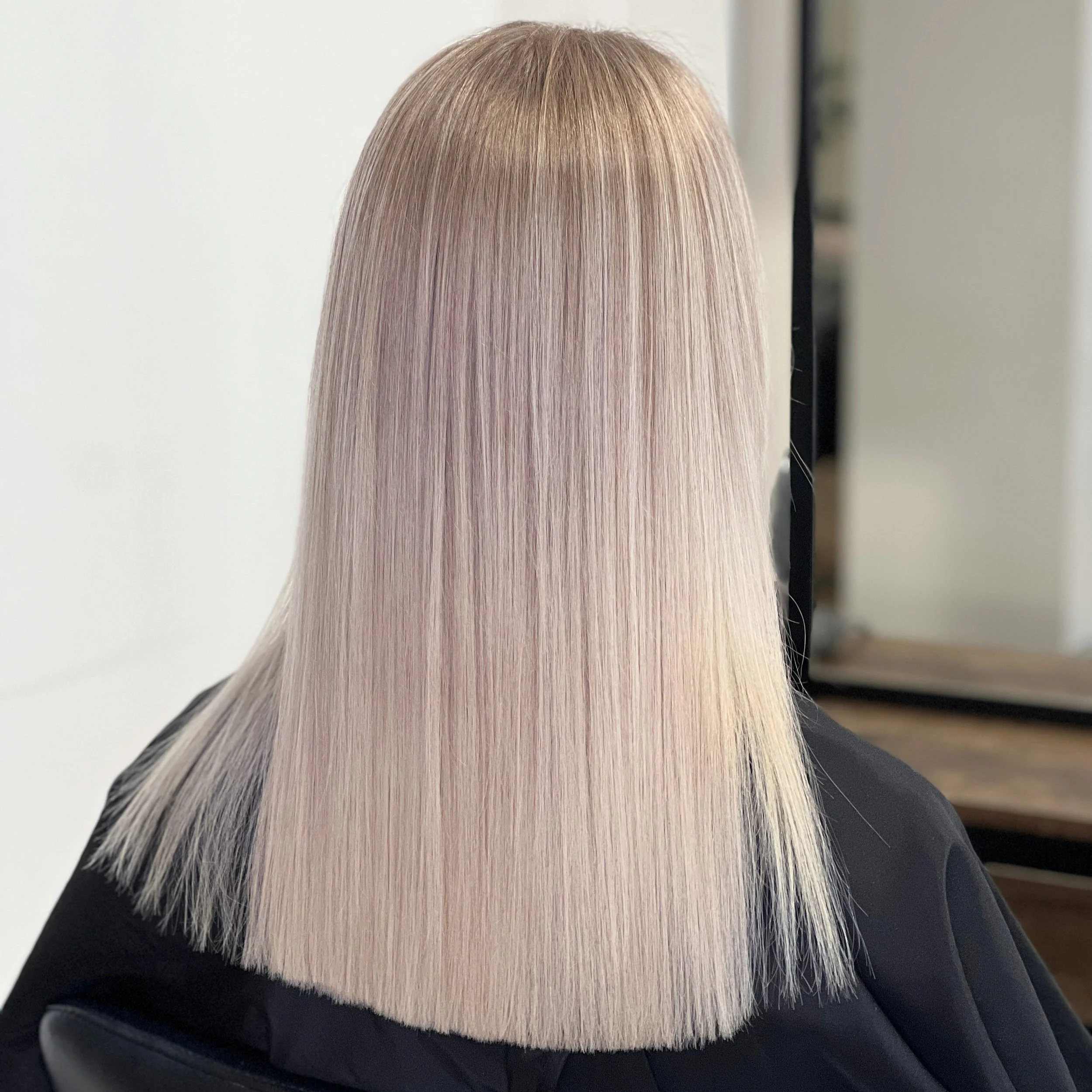 Back view of a person with straight, platinum blonde hair with a smooth, shiny finish, cut in a blunt style at the shoulders, sitting in a salon chair.