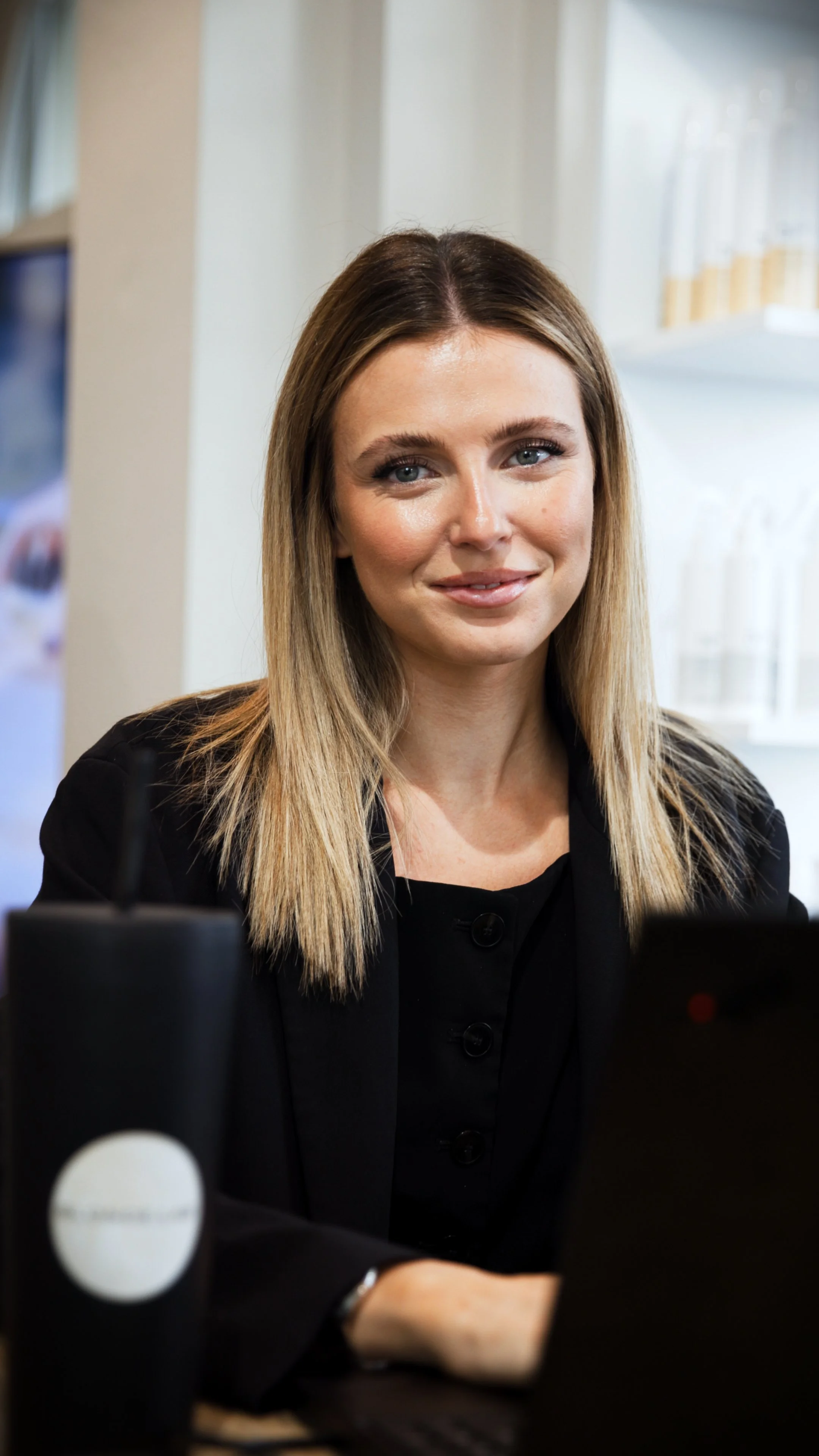 A modern hair salon featuring beige styling chairs around a white movable station with mirrors. The salon has white walls, wooden beams on the ceiling, and natural light from two windows, with a large green plant in the corner.