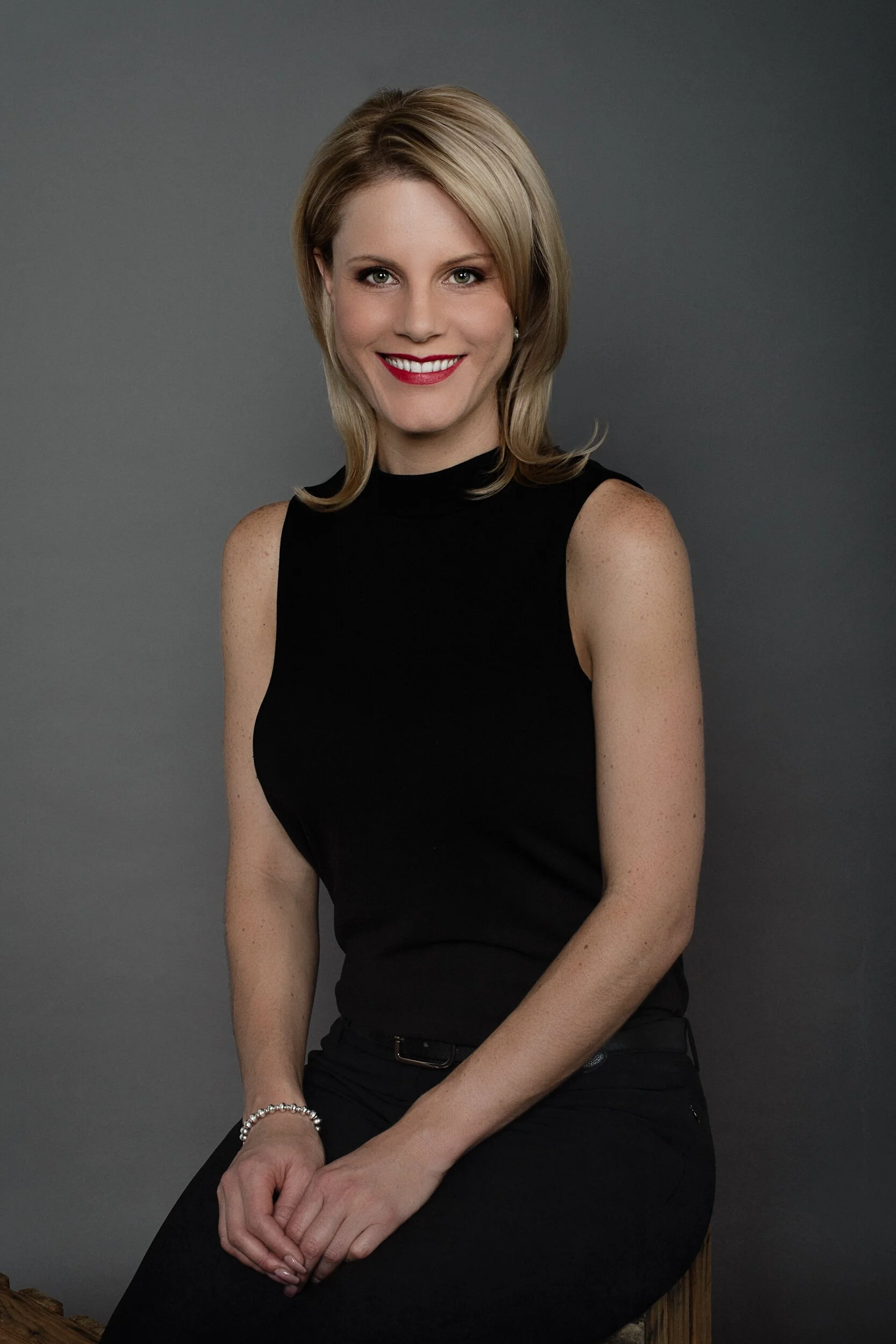 A woman with blonde hair wearing a black sleeveless top and black pants, smiling while sitting on a stool against a gray background.