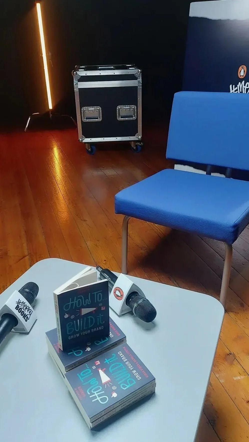 A stack of "How To Build It: Grow Your Brand" books on a grey table with two branded Penguin and #Merky Books microphones in a professional studio setting with a blue chair and stage lighting.