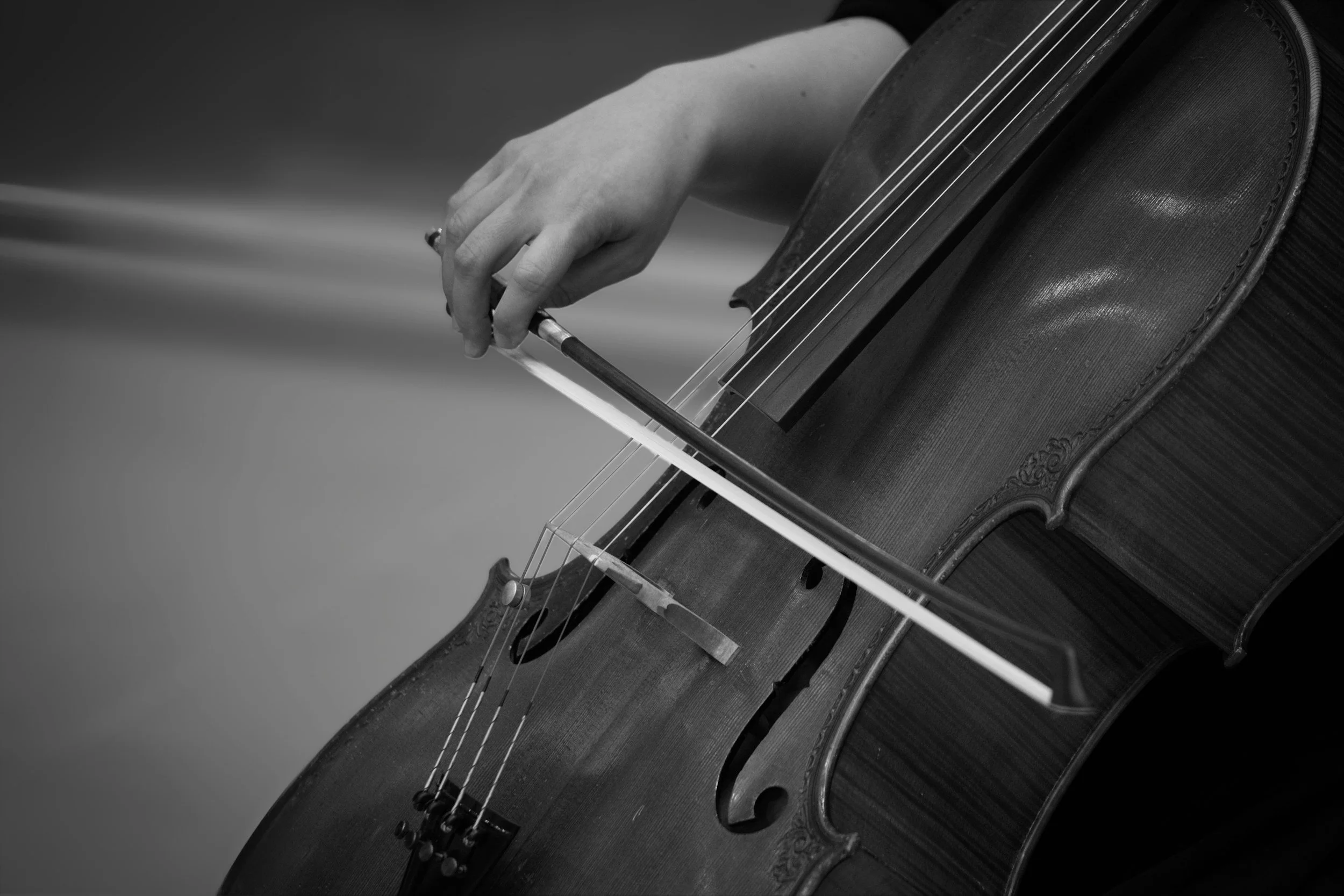 A person playing a cello, focusing on the hand holding the bow and the strings of the cello in black and white.