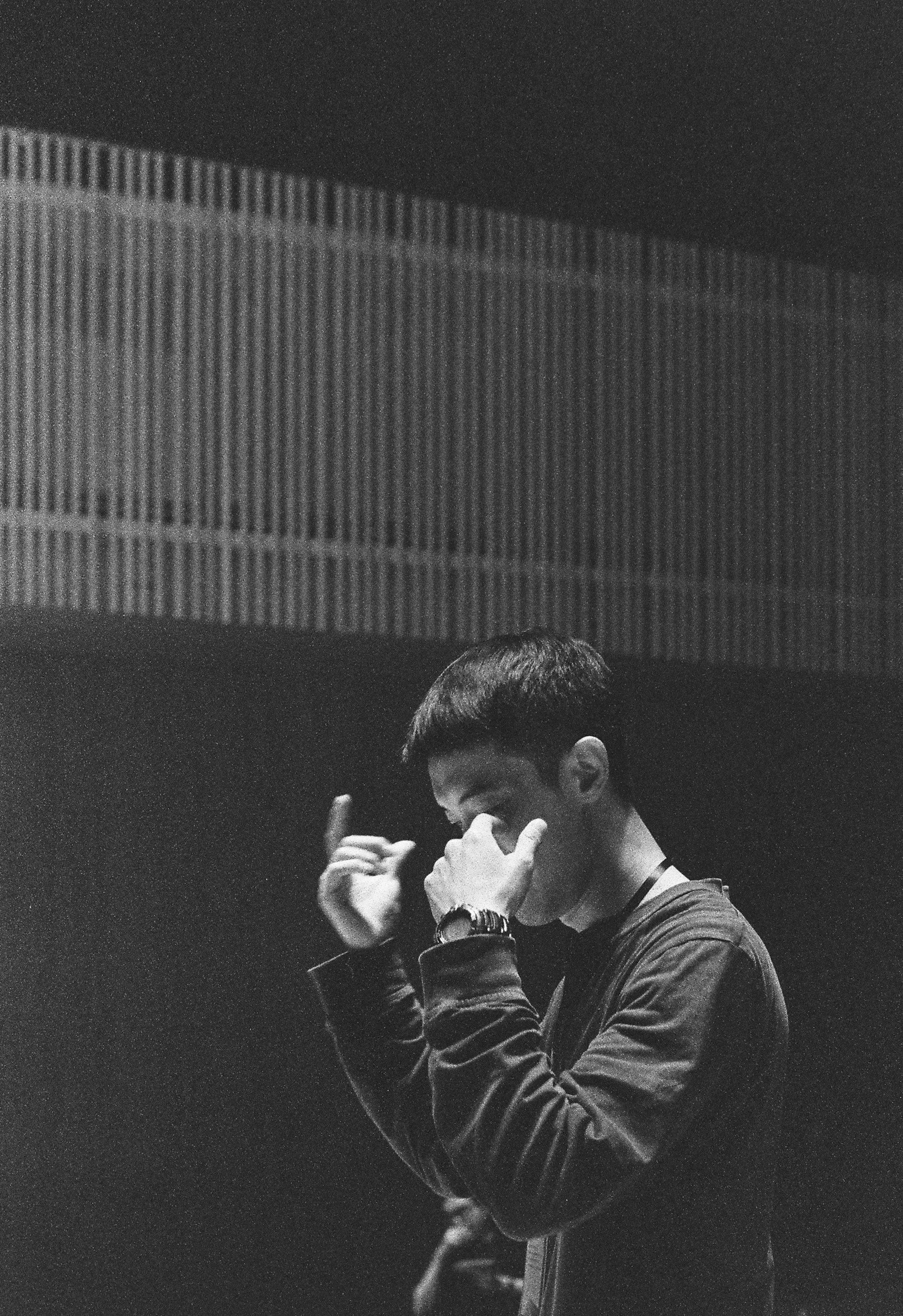 A black and white photograph of a young man with short hair, wearing a jacket, holding his nose and making a hand gesture with his index and pinky fingers extended. The background features a wall with vertical lines and a horizontal strip of pattern.