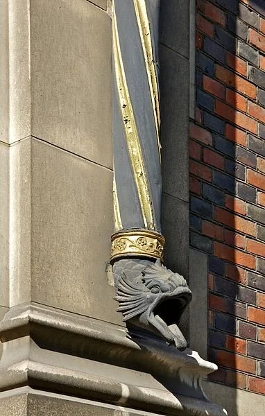 Close-up of a decorative gargoyle-style downspout on a building, featuring a dragon head with an open mouth, located at the intersection of beige stone and red brick walls.