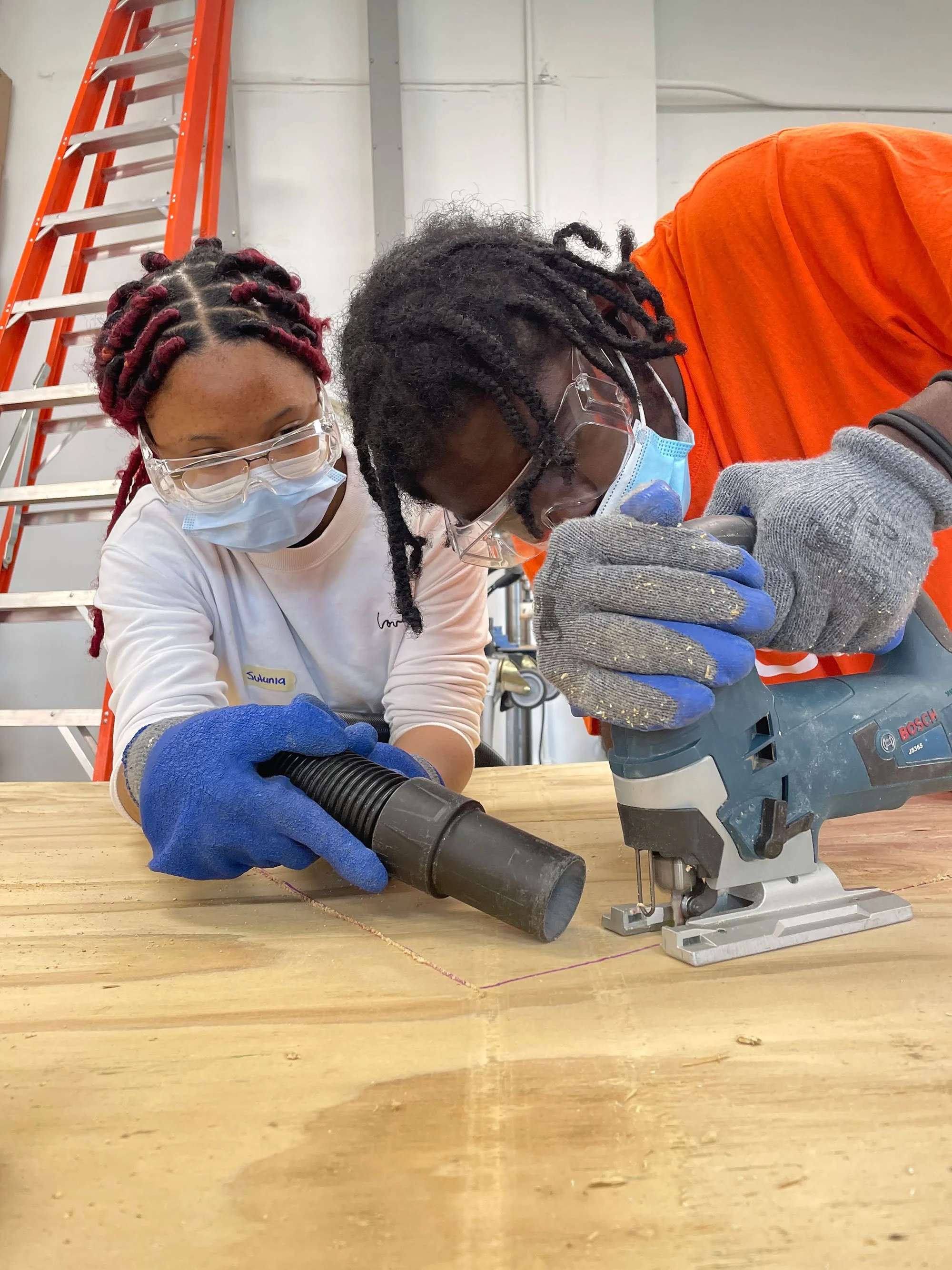 Two project fellows working together to cut wood with with a jigsaw. One is holding the saw and the other is holding a vacuum hose.