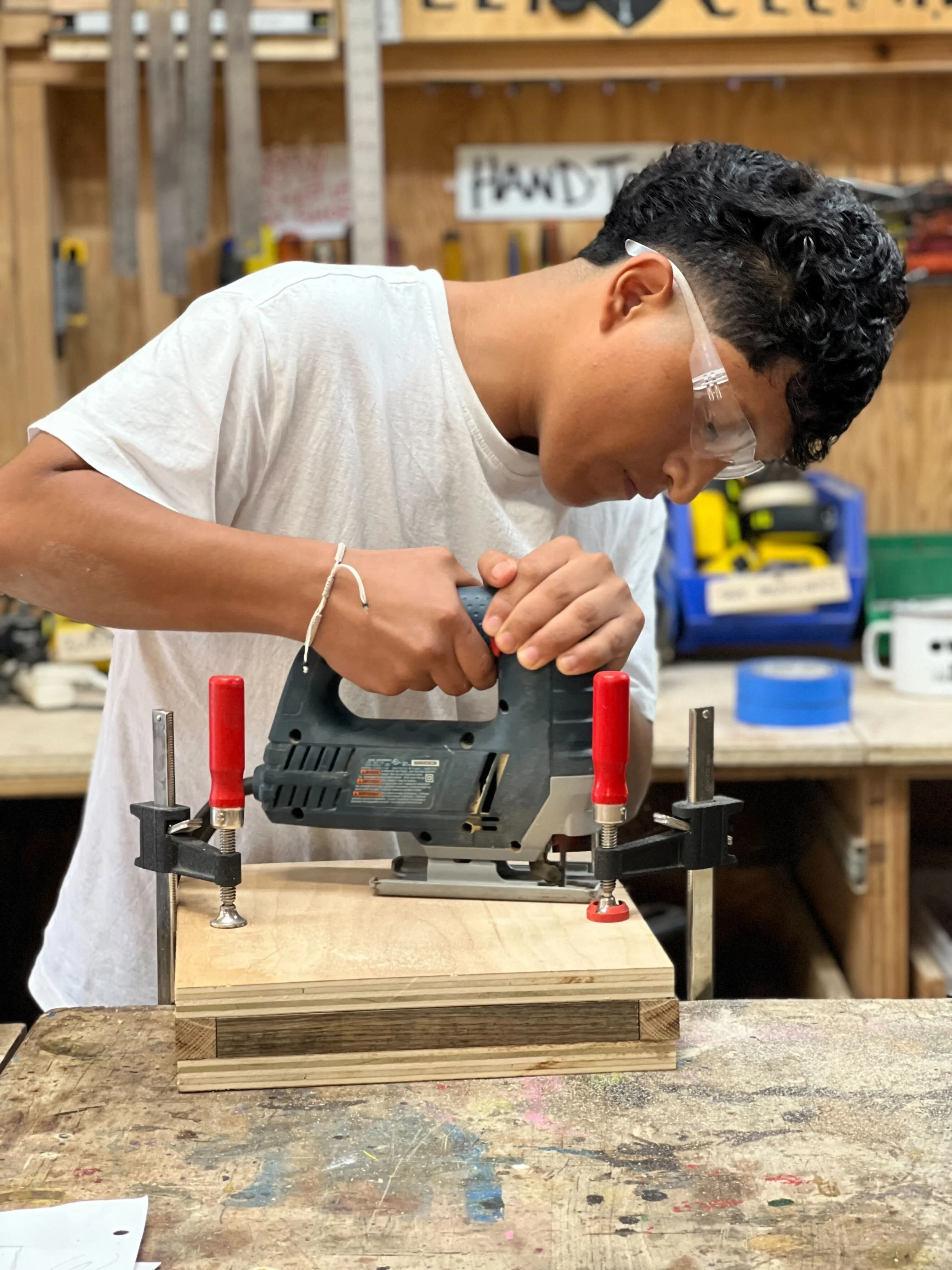A young person exploring jig saw and power tools at a woodworking station in a workshop, surrounded by various tools and equipment.