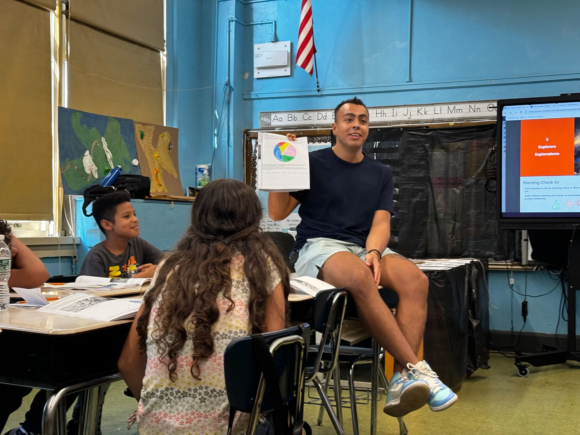 ELL teacher in classroom with students sitting on a desk and talking while holding up a workbook