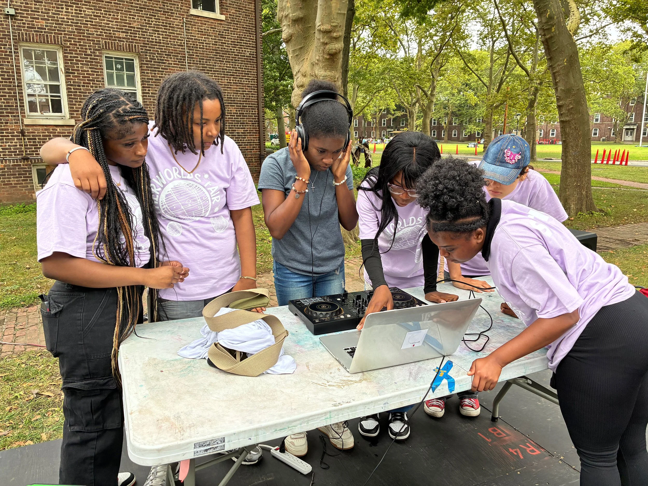 A group of students at Beam Center's OtherWorlds Fair gathered around a computer on a table with a DJ system listening to headphones and adjusting something on the computer