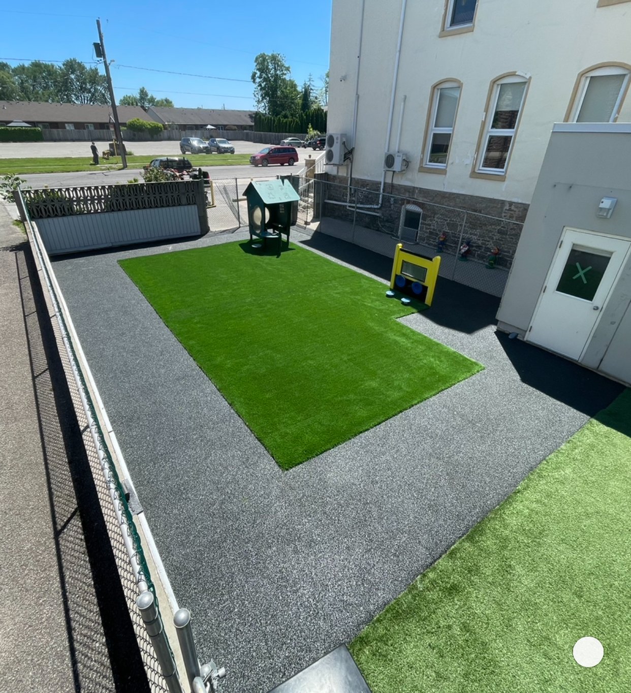 Outdoor playground area with artificial grass, small playhouse, and interactive panels, surrounded by fence.