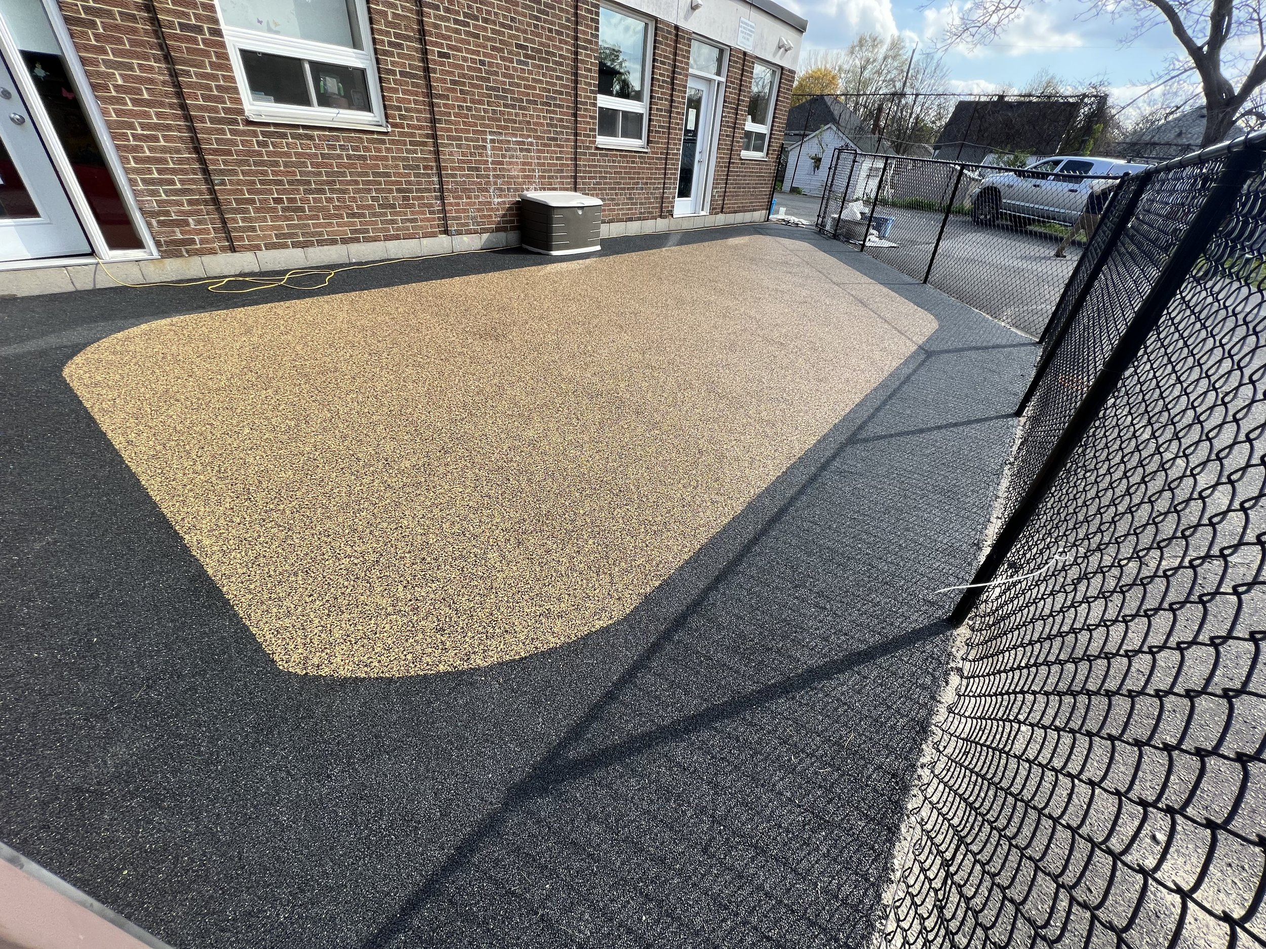 Fenced outdoor area with a textured rubber flooring in black and tan, adjacent to a brick building with windows and doors.