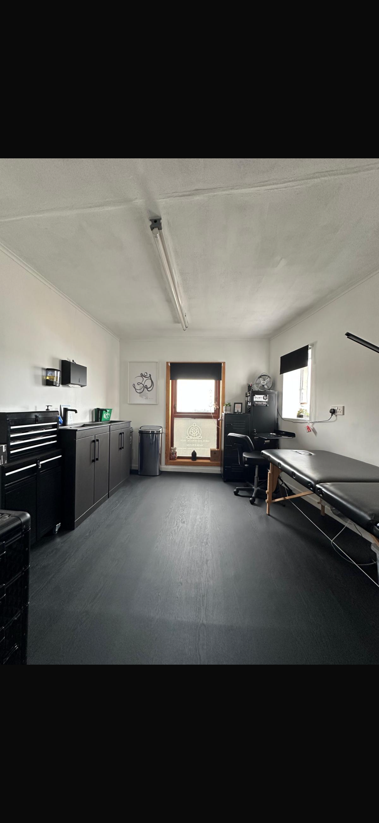 A room with a massage table, a desk with a computer, cabinets, and artwork, illuminated by natural light from two windows.
