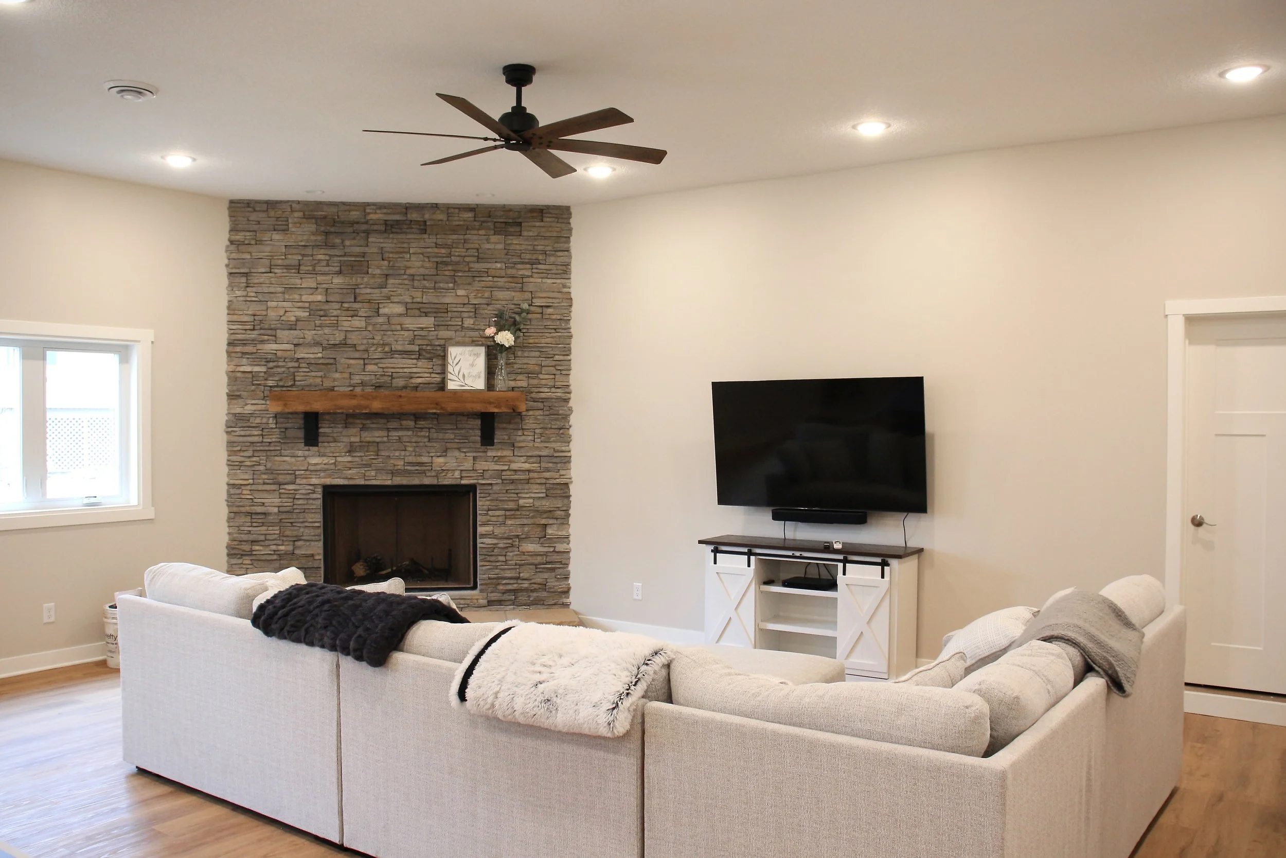 Living room with a beige sofa facing a wall-mounted TV and a stone fireplace with wooden mantel.