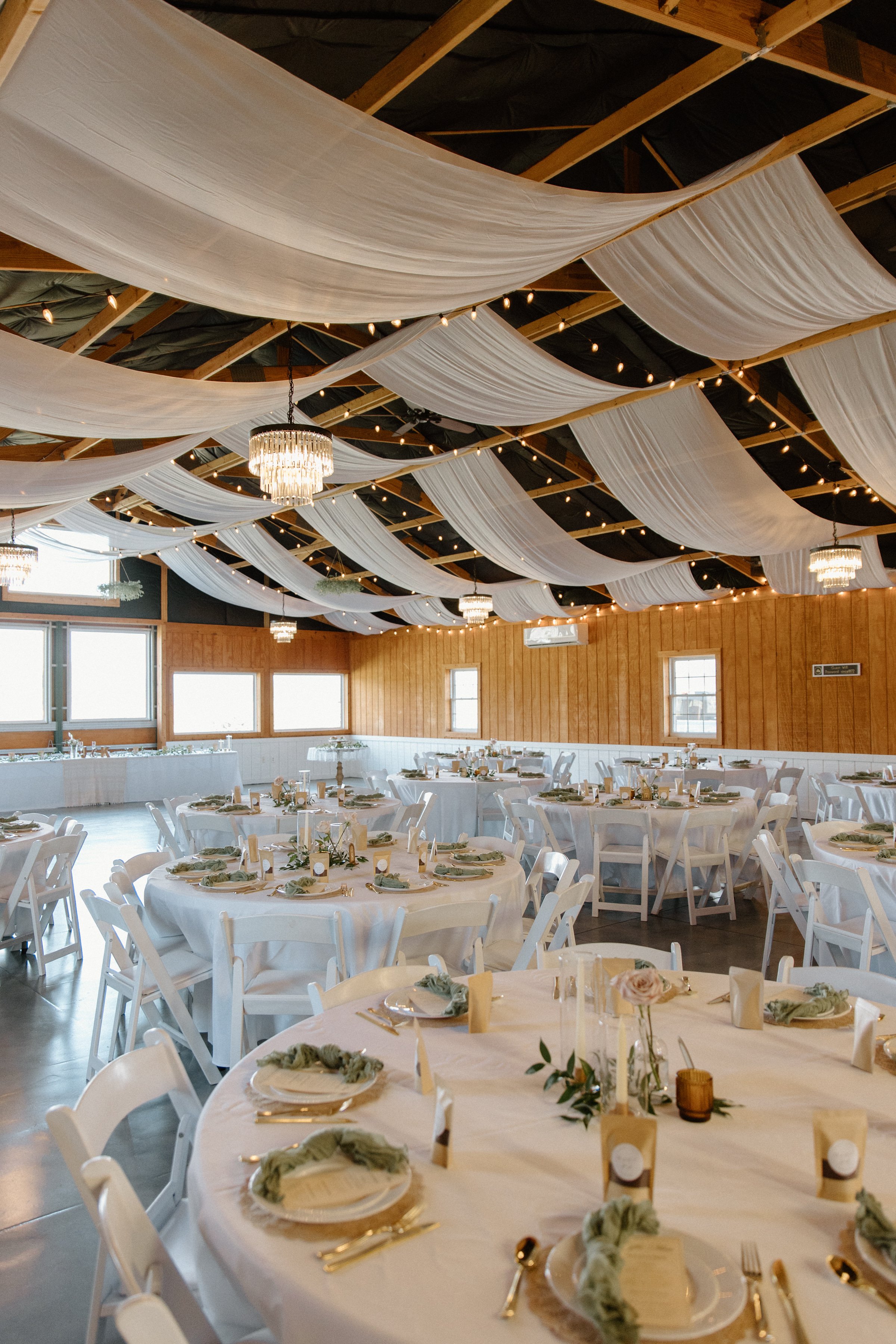 Decorated event hall with round tables covered in white tablecloths, set with plates, gold utensils, cloth napkins, candles, and floral centerpieces. White chairs surround the tables. The ceiling features white fabric drapes and string lights, with s