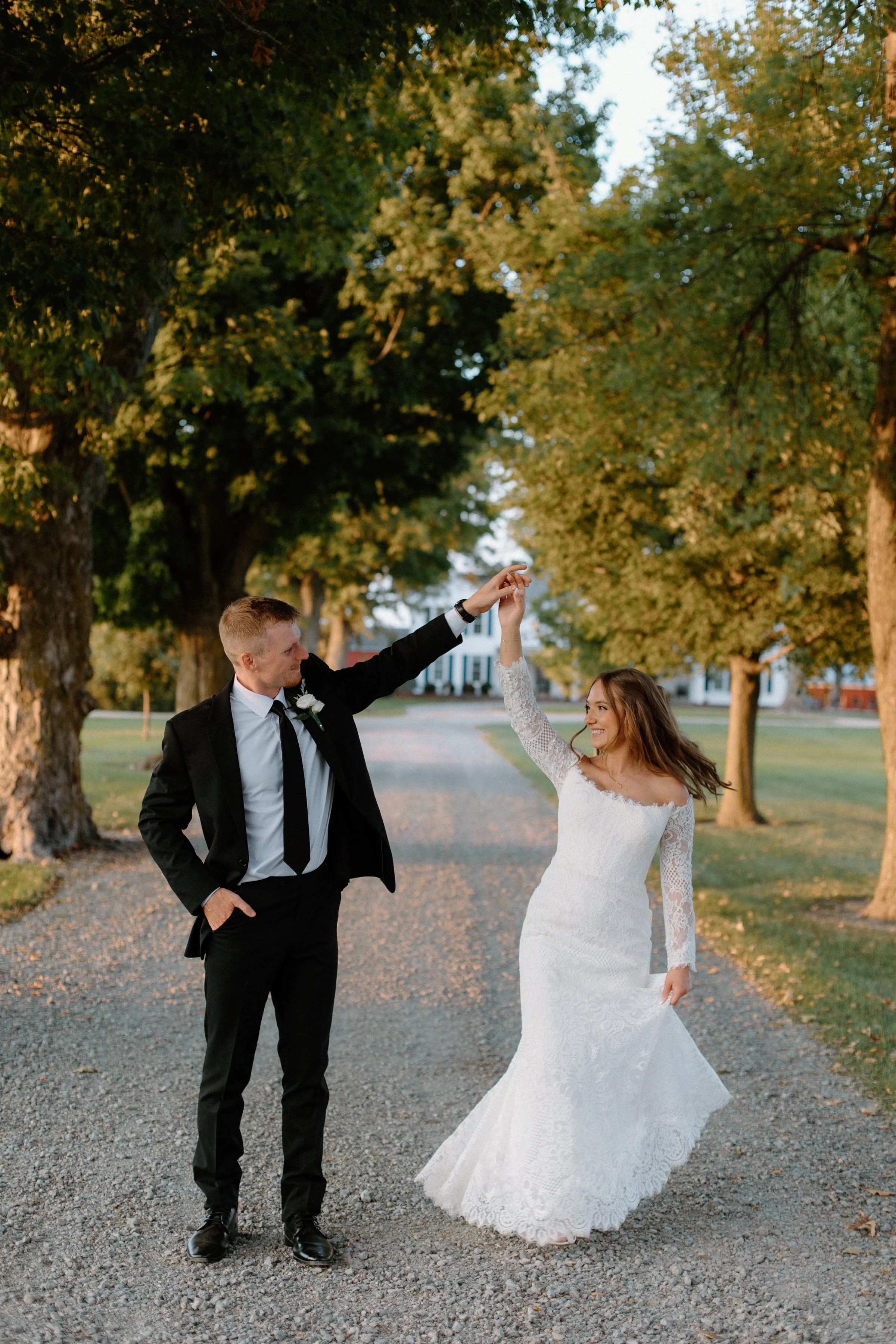 A bride and groom dancing outdoors on a gravel pathway surrounded by trees during sunset.