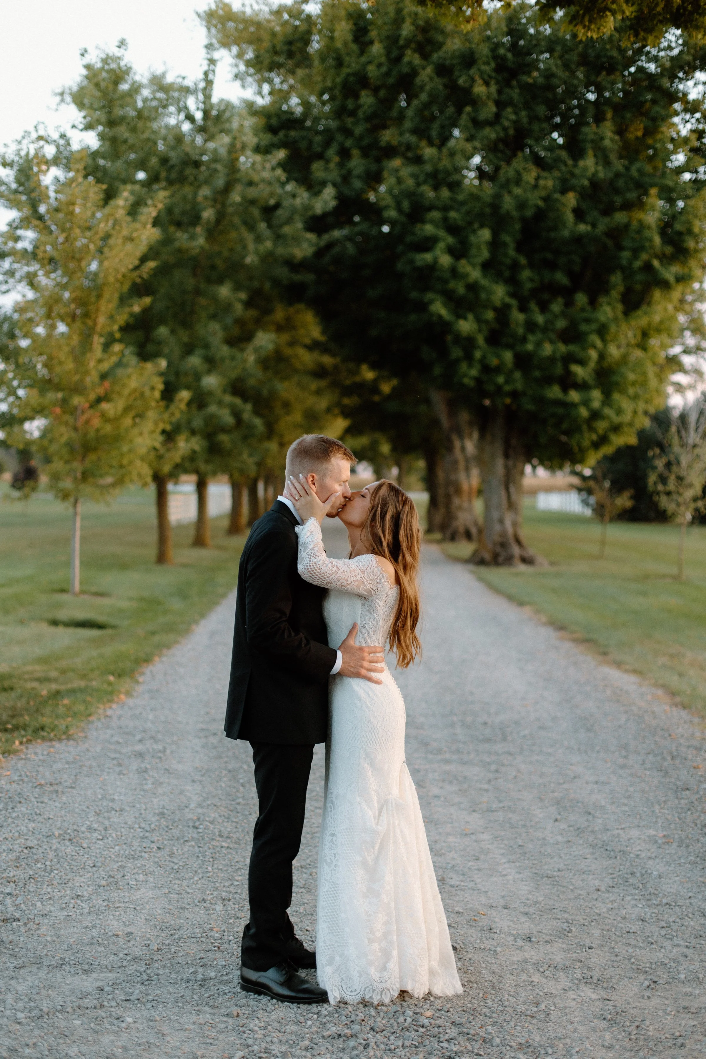A bride and groom share a kiss on a gravel path lined with green trees during sunset.