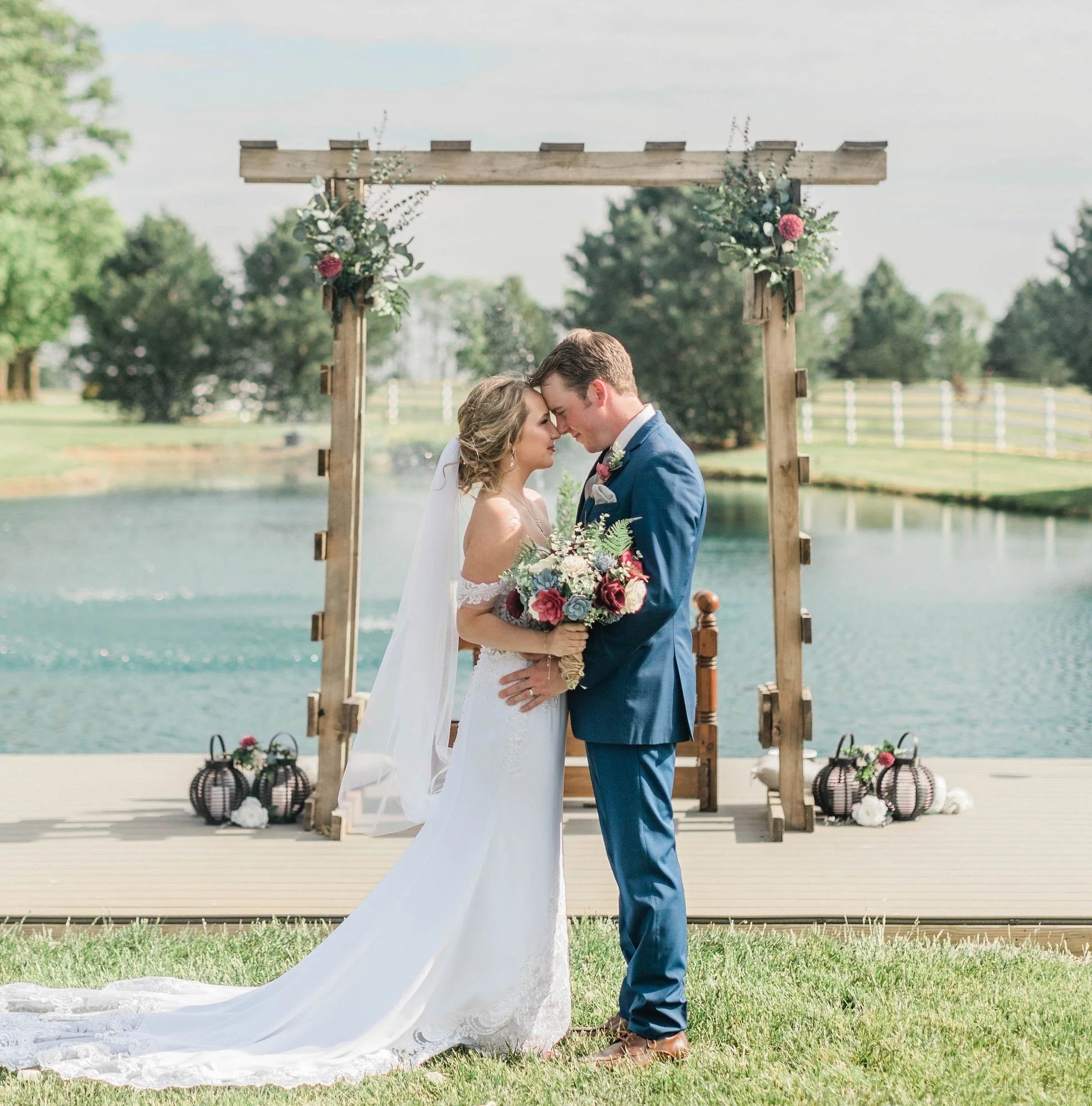 A bride and groom standing close together outdoors by a lake during their wedding, touching foreheads under a wooden arch decorated with flowers, with lanterns and white flowers on the ground nearby.