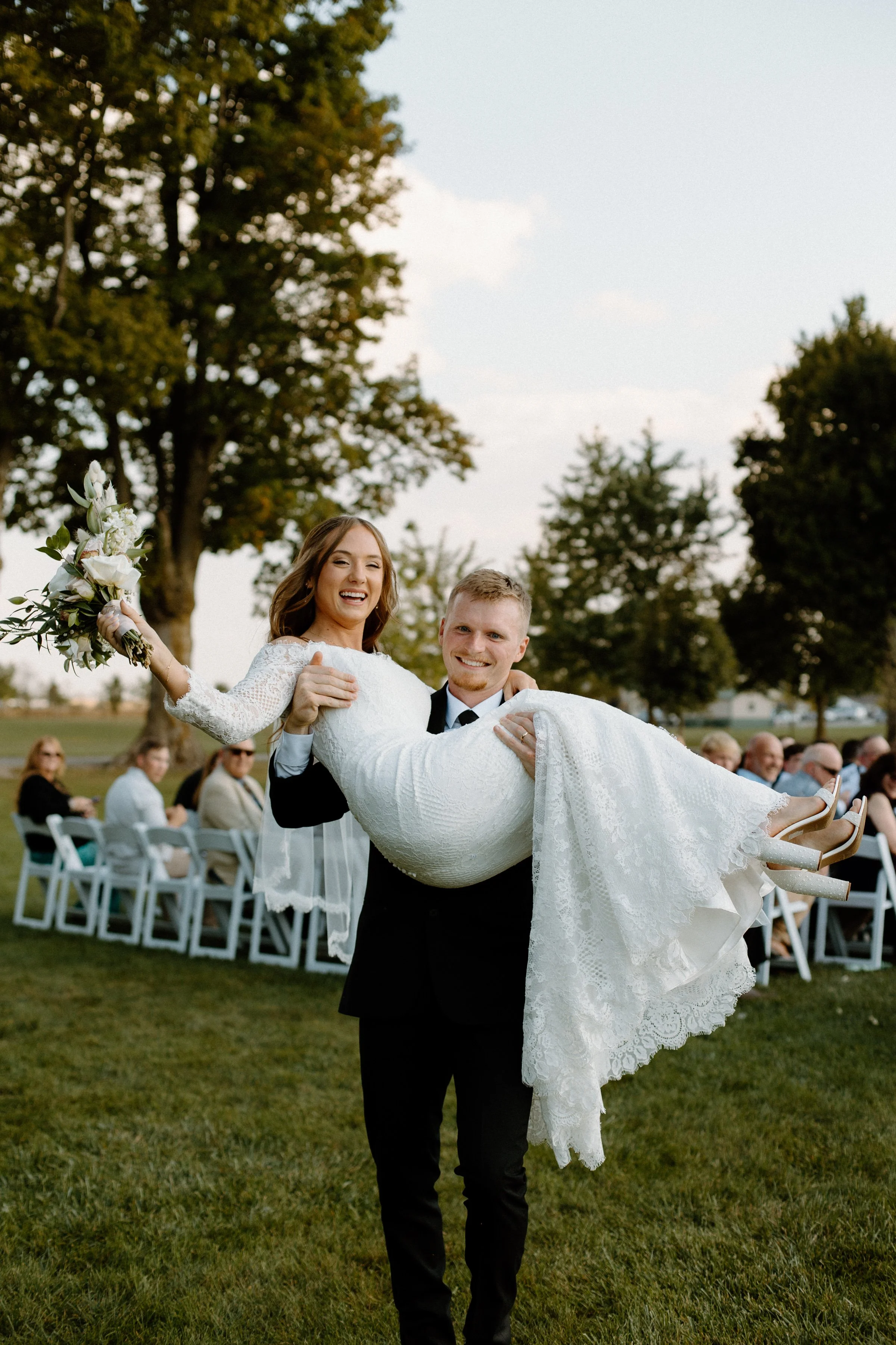 A groom in a black suit carrying a smiling bride in a lace wedding dress outdoors during a wedding ceremony, with guests seated in the background and trees under a partly cloudy sky.