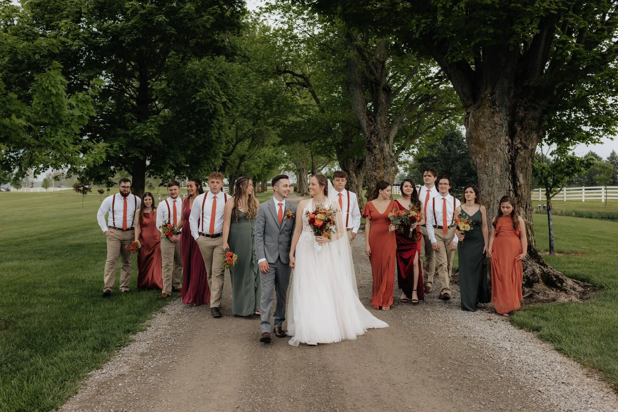 A wedding party of 14 people walking outdoors on a gravel path surrounded by green trees and grass, with the bride and groom in the center. The bride is dressed in a white gown holding a bouquet, and the groom is in a gray suit with a orange tie. The