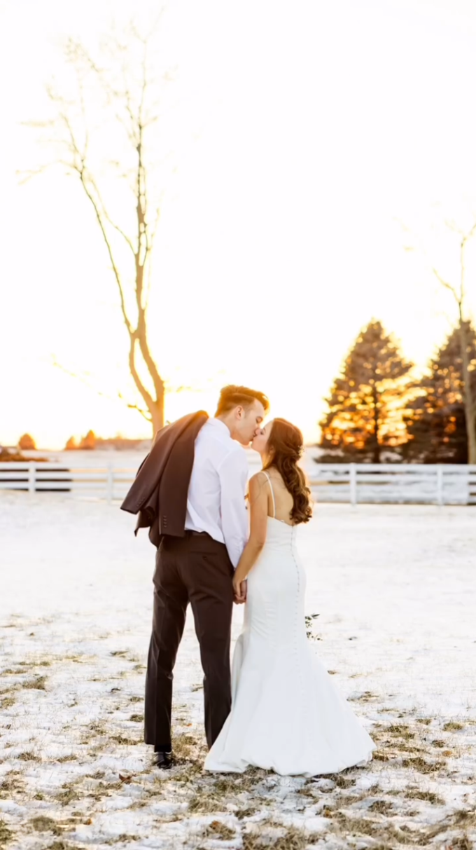 A bride and groom sharing a kiss outdoors during sunset, with leafless trees and a white fence in the background.