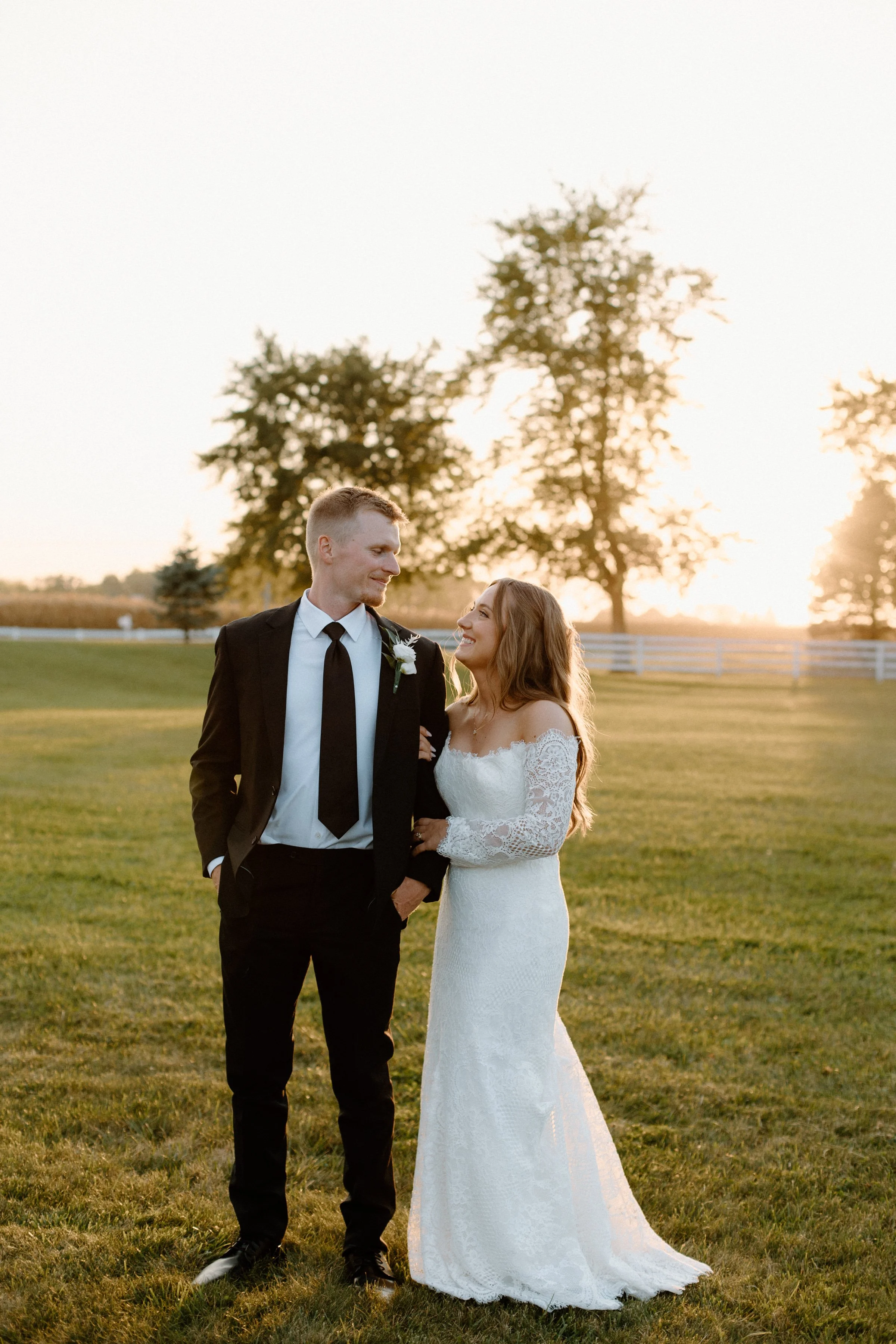 A newlywed couple standing on a grassy field during sunset. The groom is wearing a black suit with a white shirt and black tie, and the bride is in a white lace wedding dress with off-the-shoulder sleeves. They are smiling and looking at each other, 
