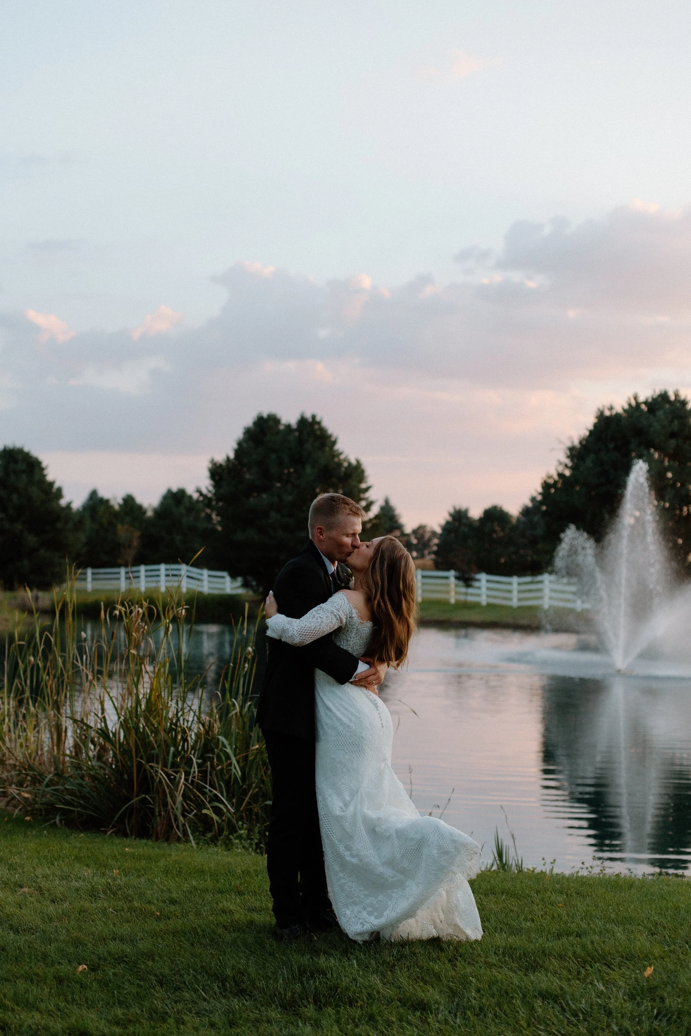 A newlywed couple sharing a kiss near a pond with a fountain at sunset, surrounded by trees and a white fence.