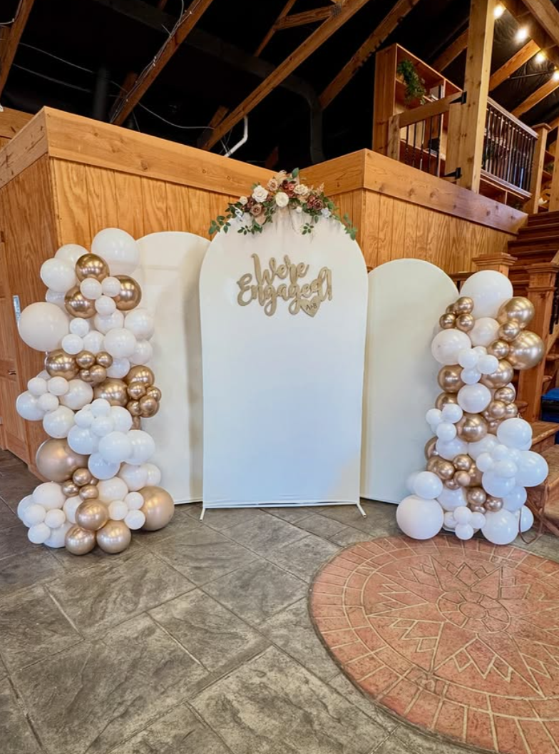 Decorated event space with round tables covered in green tablecloths, set for a celebration, with floral centerpieces, candles, and place settings. The ceiling is draped with white fabric and string lights, and there is a balloon arch with pastel-colored balloons and a sign that reads 'Oh Baby' on the left side of the room. Large windows show a scenic outdoor view.