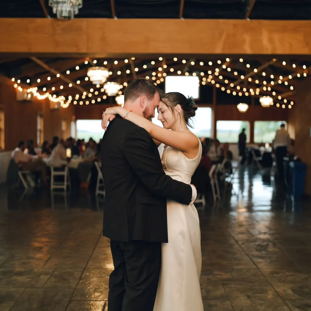A bride and groom share a dance at their wedding reception in a decorated hall with string lights and hanging chandeliers, surrounded by seated guests.