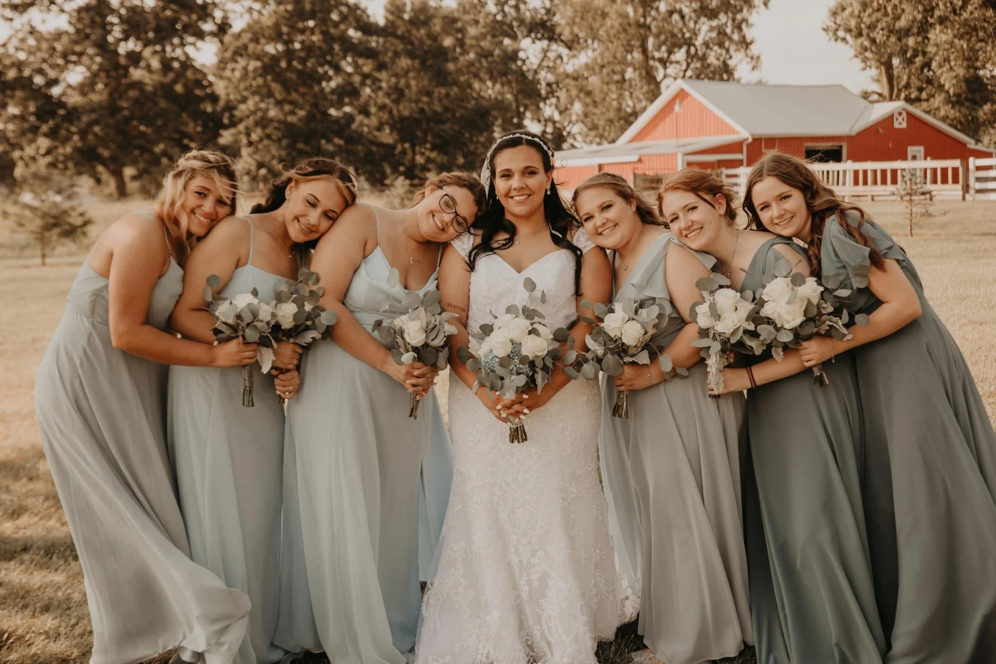 A bride and six bridesmaids at an outdoor wedding, all holding bouquets of white and green flowers, with trees and a red barn in the background.