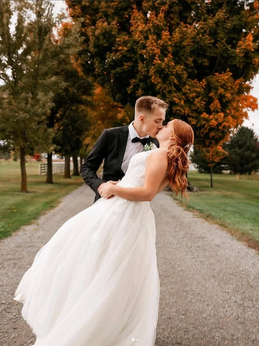 A groom in a black tuxedo and a bride in a white wedding dress sharing a kiss outdoors on a gravel path, with trees in autumn colors in the background.