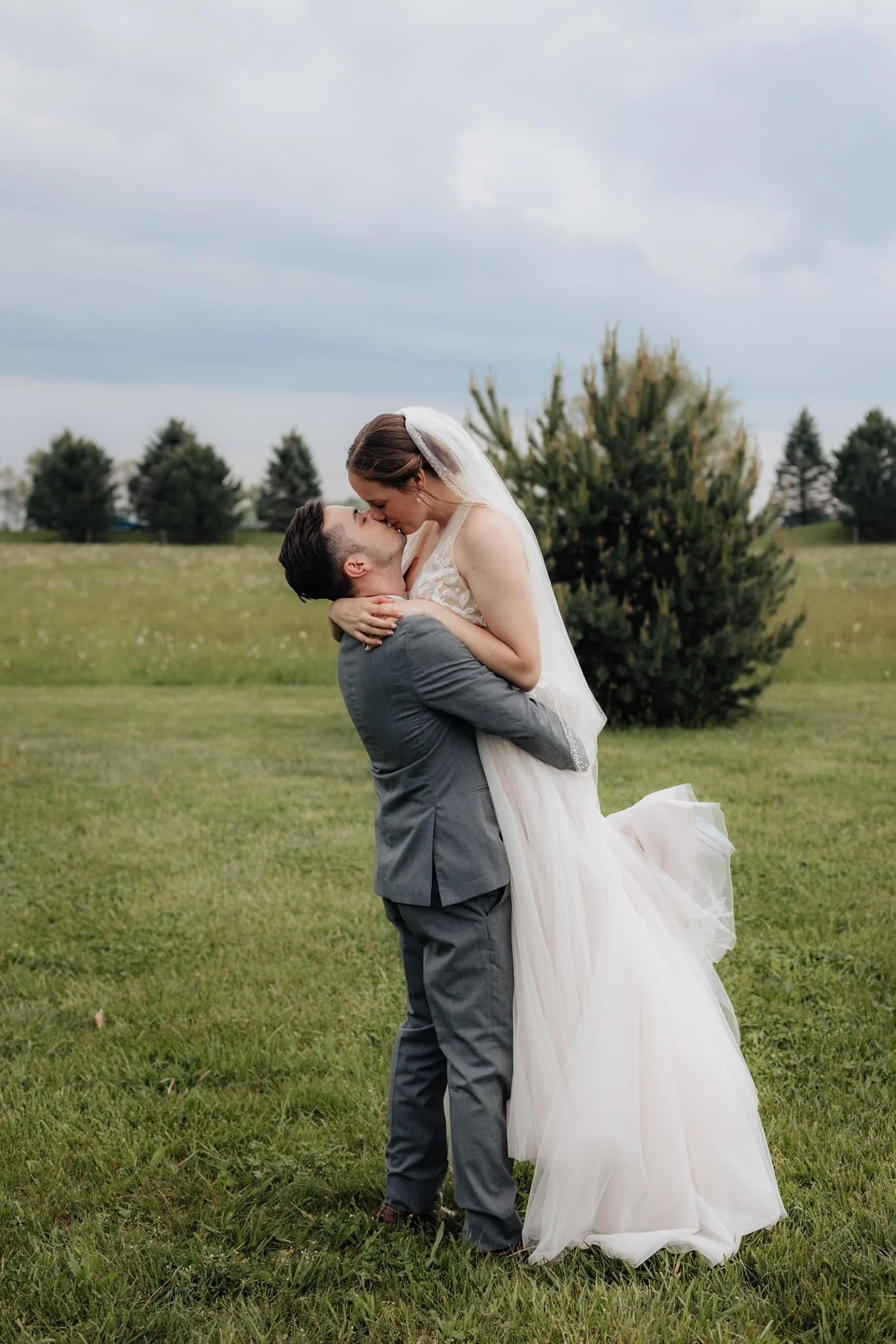 A bride and groom sharing a kiss outdoors in a grassy field, with trees and a cloudy sky in the background.