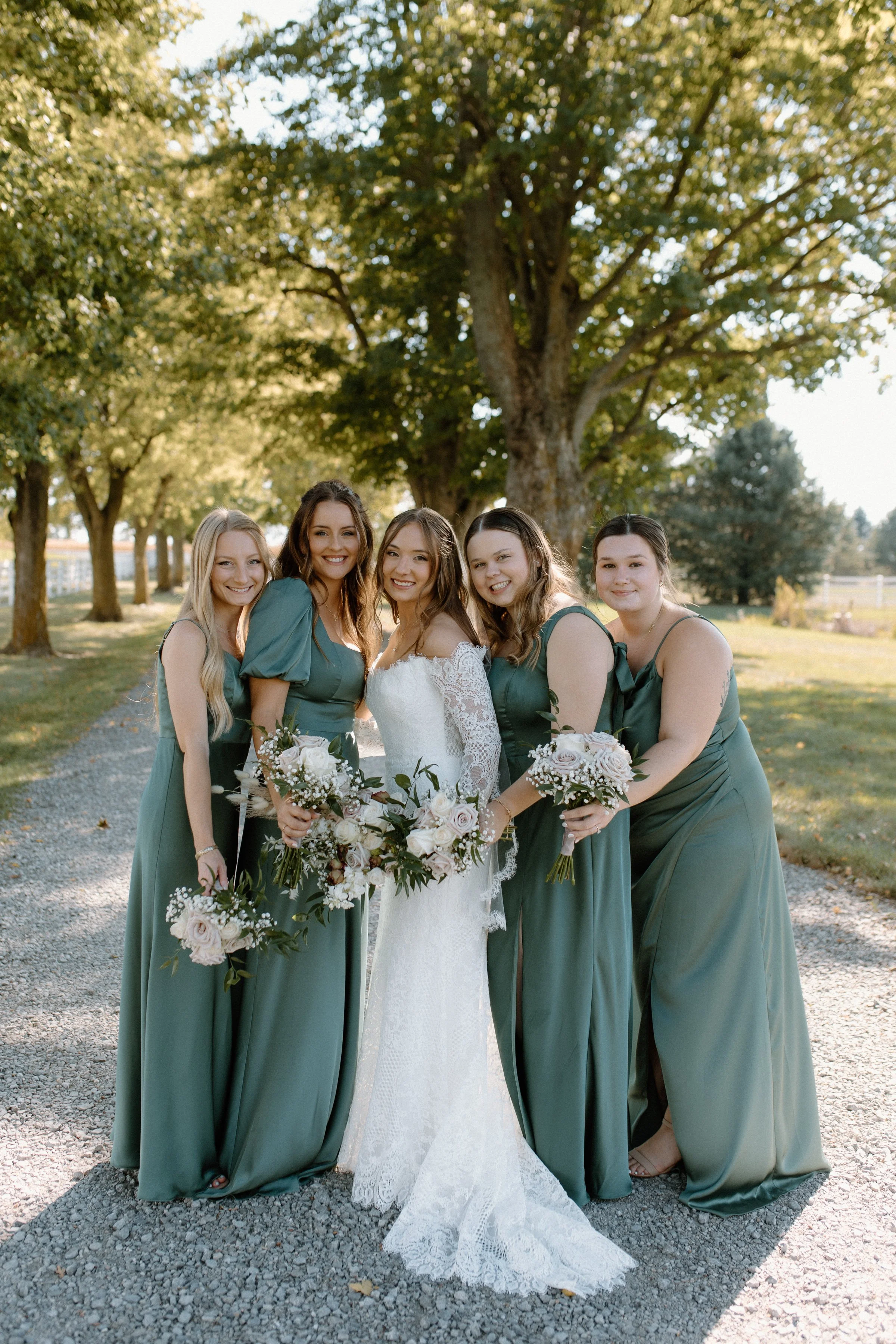 A bride and four bridesmaids posing outdoors on a gravel path surrounded by trees, with sunlight filtering through the leaves. The bride is wearing a white lace wedding gown, and the bridesmaids are in matching dark green dresses. They are holding bo