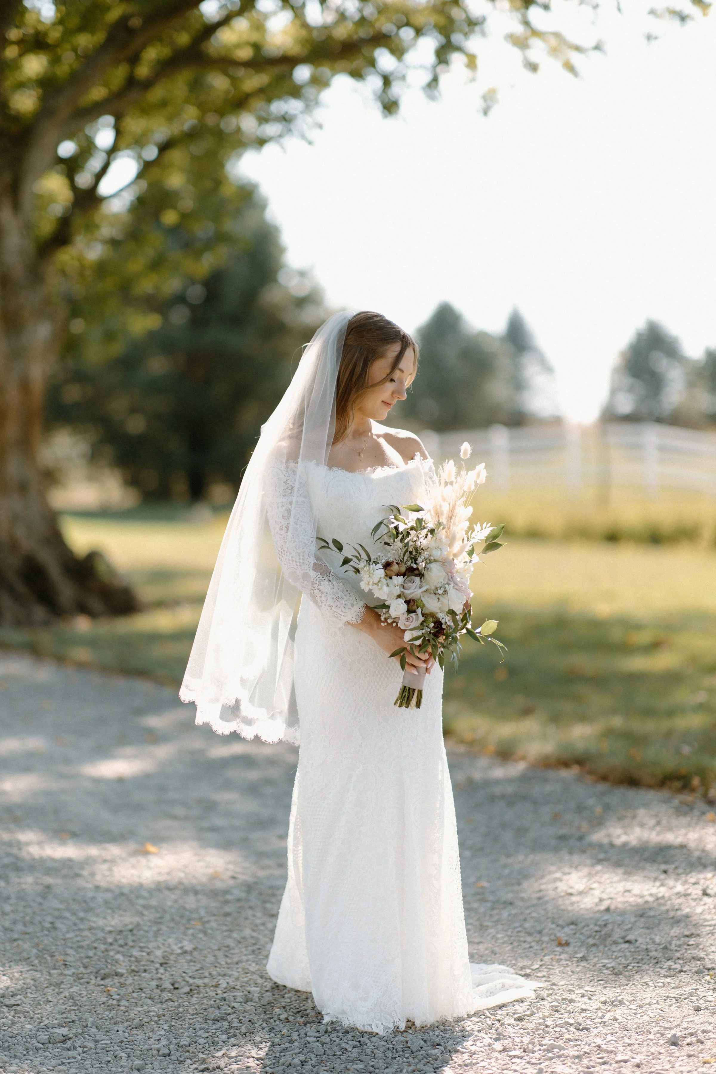 A bride in a white wedding dress holding a bouquet, standing outdoors on a gravel path with trees in the background.