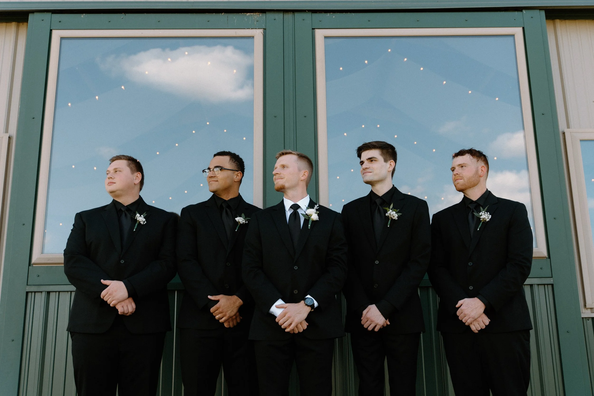 Five men dressed in black suits and ties standing in front of a building with large windows reflecting a blue sky with clouds. They are wearing boutonnières on their lapels and are posed with their hands clasped.