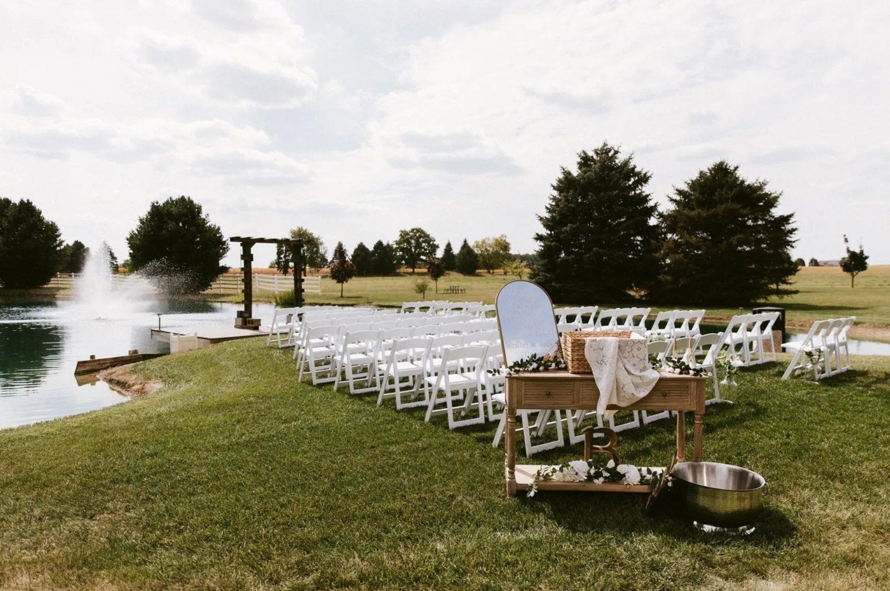 Outdoor wedding setup beside a pond with rows of white chairs, a table with a mirror, flowers, a basket, and a dog bowl, and a fountain in the background under a cloudy sky.