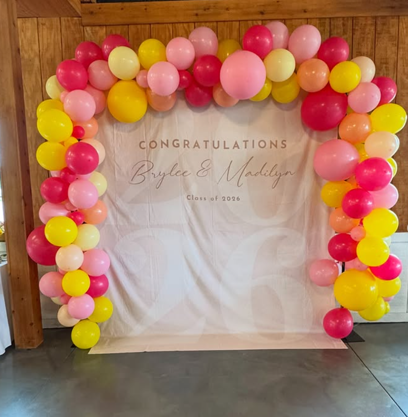 Indoor celebration hall decorated with white drapery and string lights for a 50th birthday party, featuring round tables with white tablecloths, floral centerpieces, candles, and black and gold balloons.