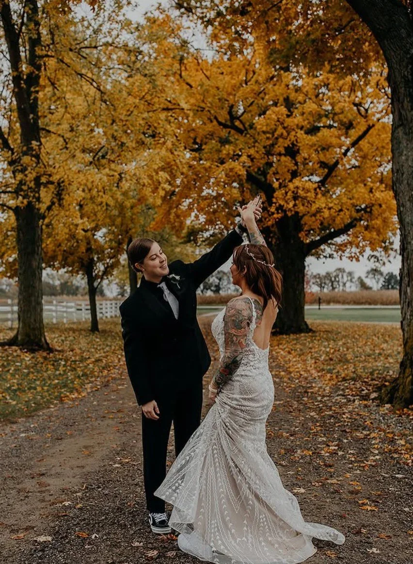 A same-sex couple dancing outdoors under autumn trees with orange leaves, one in a black suit and the other in a white wedding gown, celebrating their wedding.