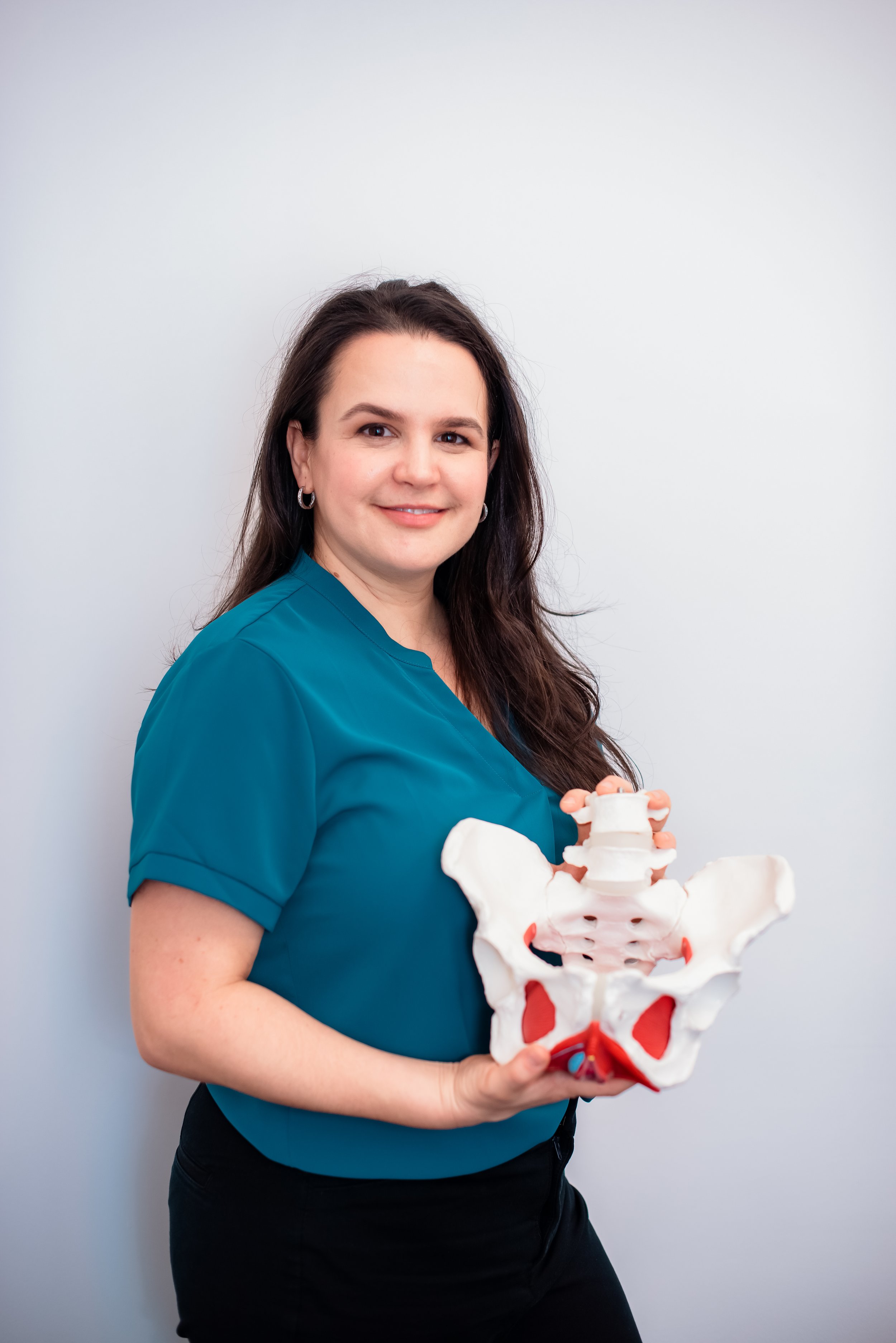 A woman with long dark hair in a teal top holding a large anatomical model of a human pelvis and lower spine, standing against a plain light-colored background.