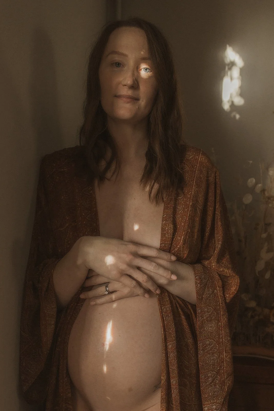 A woman with red hair standing next to a wall, wearing a brown patterned robe open to reveal her bare chest and stomach, with light spots on her skin and a ring on her finger.