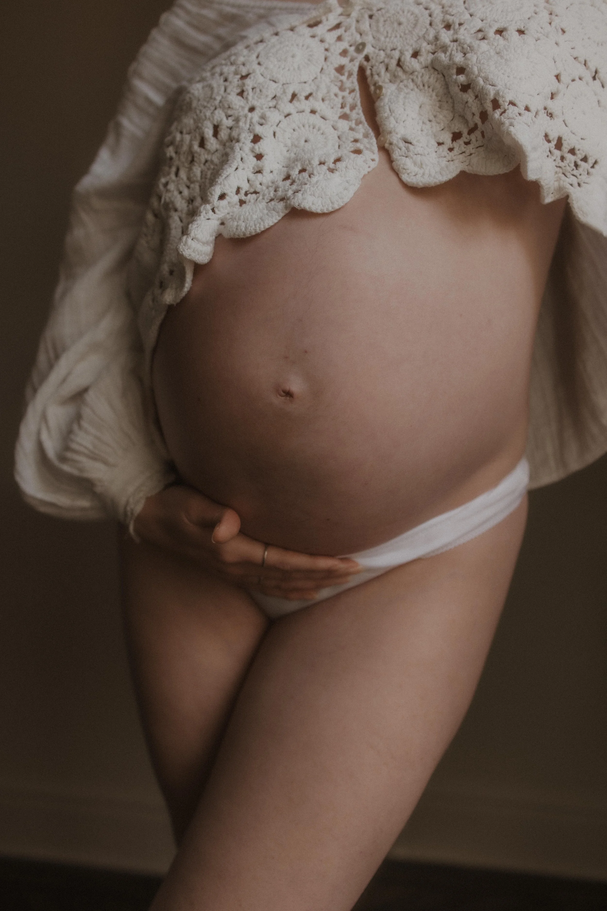Close-up of a pregnant woman's belly with one hand resting on her lower abdomen, wearing a cream-colored knitted sweater and white underwear.