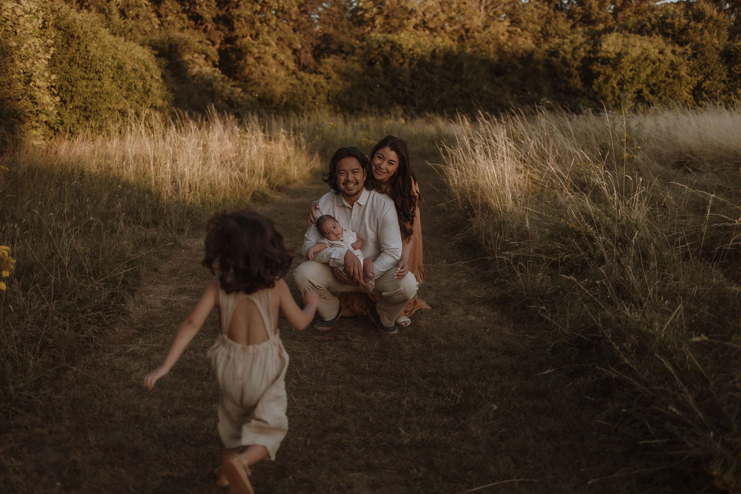 A family of four enjoying a walk in a natural setting during sunset, with the parents kneeling and the children running on a dirt path surrounded by tall grass and trees.