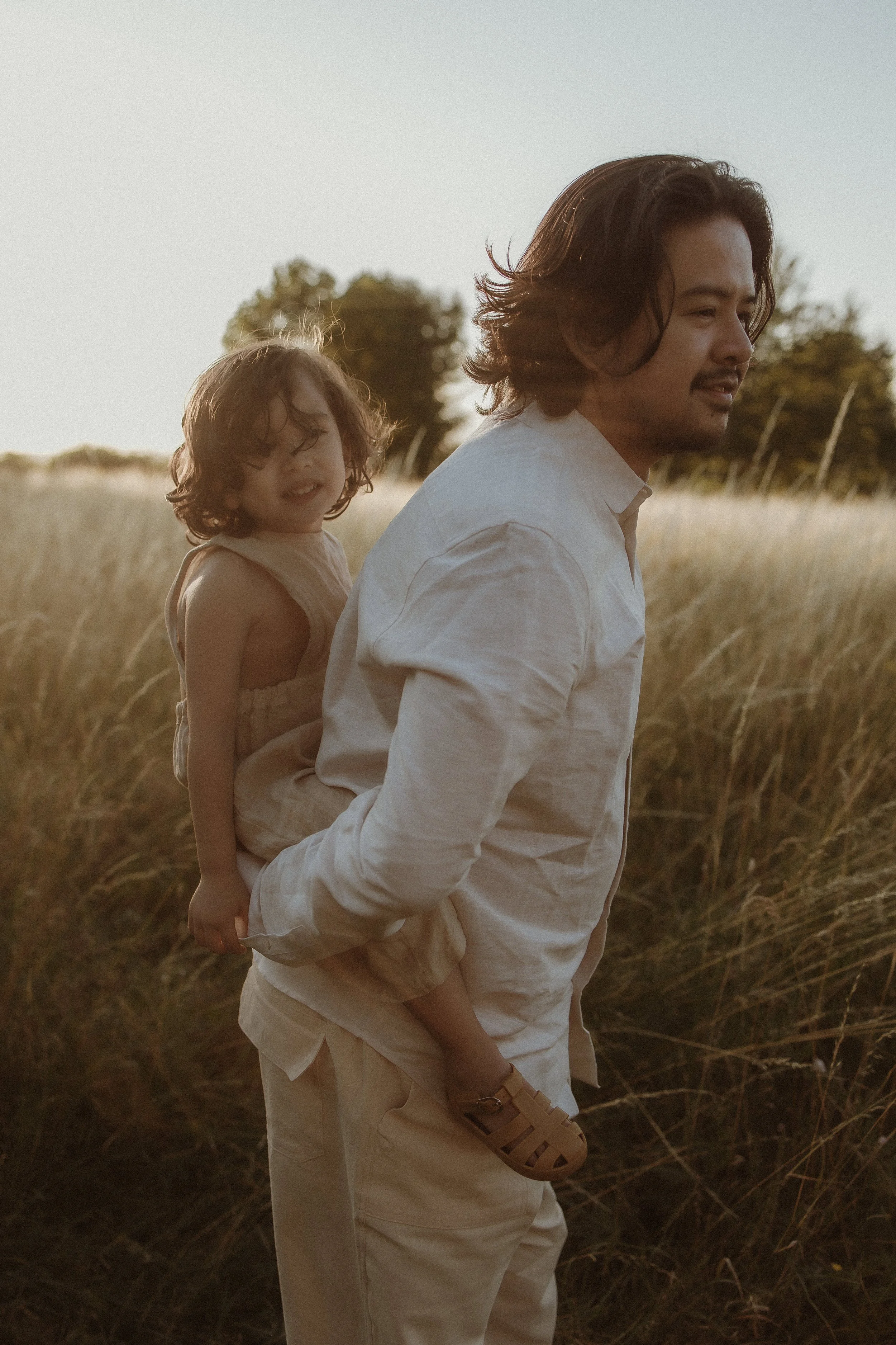 A man walking in a field with a young girl on his back, both dressed in light-colored clothing, during sunset.