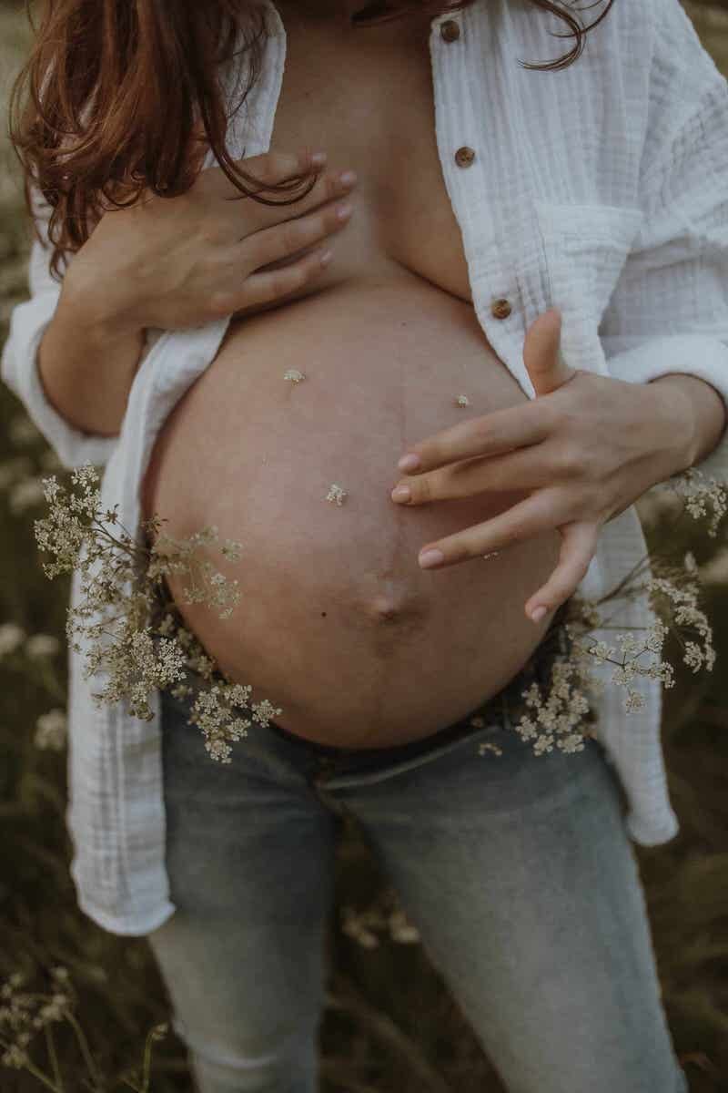 Pregnant woman outdoors wearing a white shirt and gray jeans, with her hands on her stomach, surrounded by small white flowers.