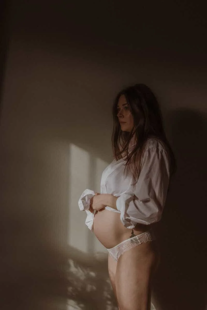 A pregnant woman standing indoors, wearing a white shirt and lace underwear, with her hand on her belly and looking to the side, illuminated by soft natural light.