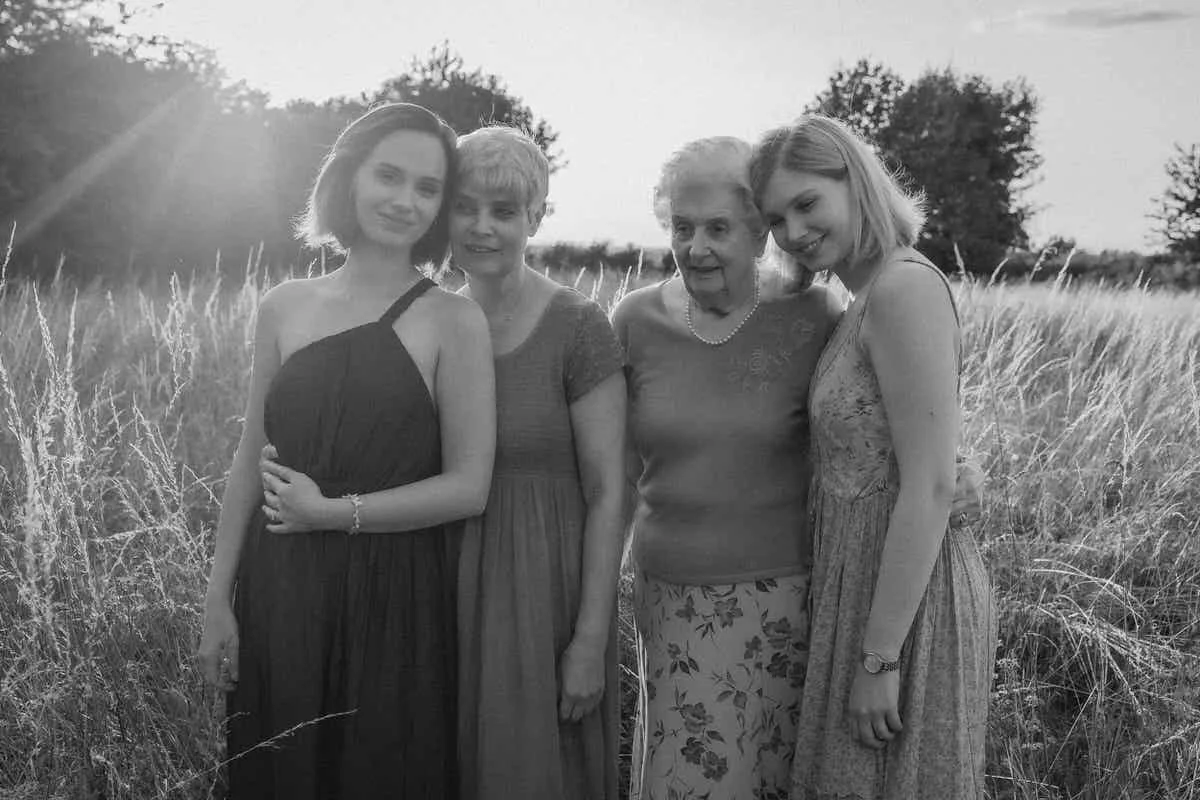 Four women standing together in a field of tall grass on a sunny day, smiling at the camera.