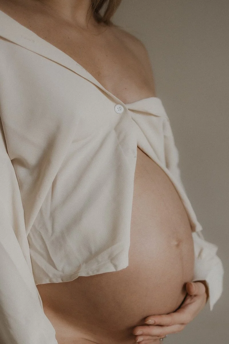 Close-up of a pregnant woman's bare belly with a hand resting on it, partially covered by an open, cream-colored shirt.