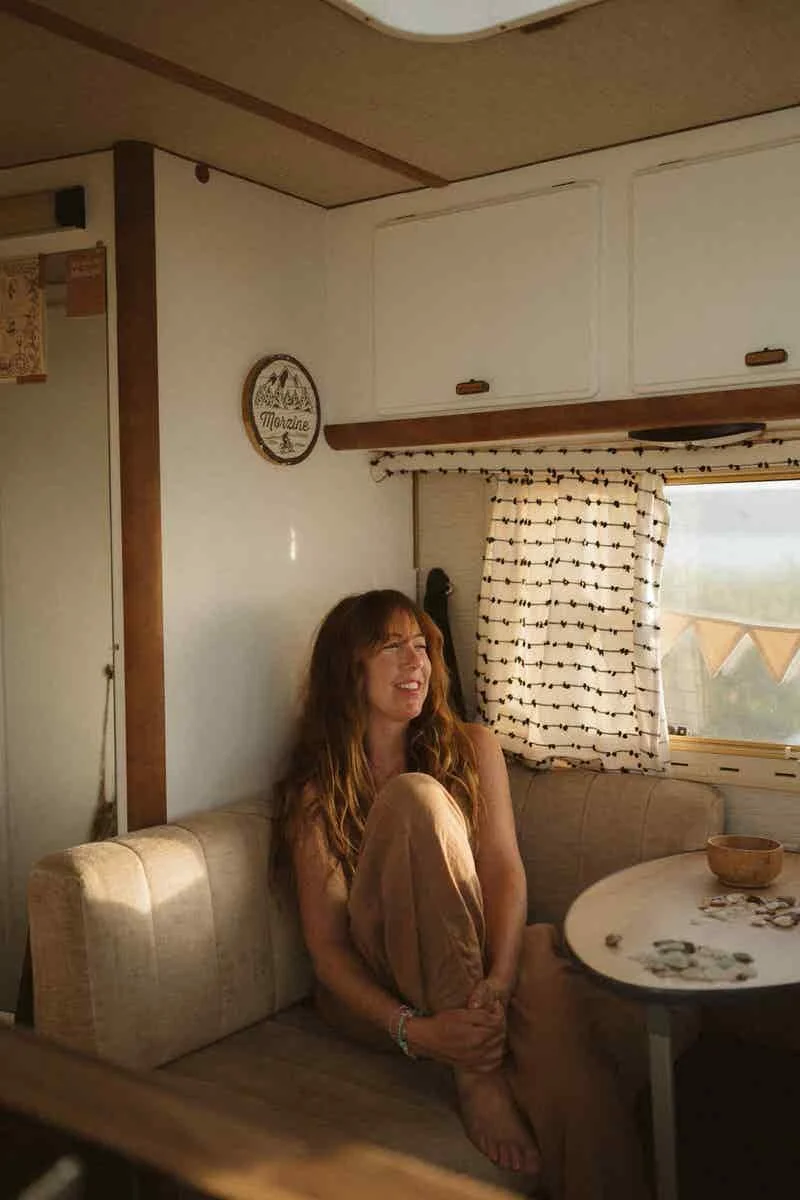 A woman sitting on a beige sofa inside an RV with a window and patterned curtain behind her, and a round table with coins and a wooden bowl in front of her.