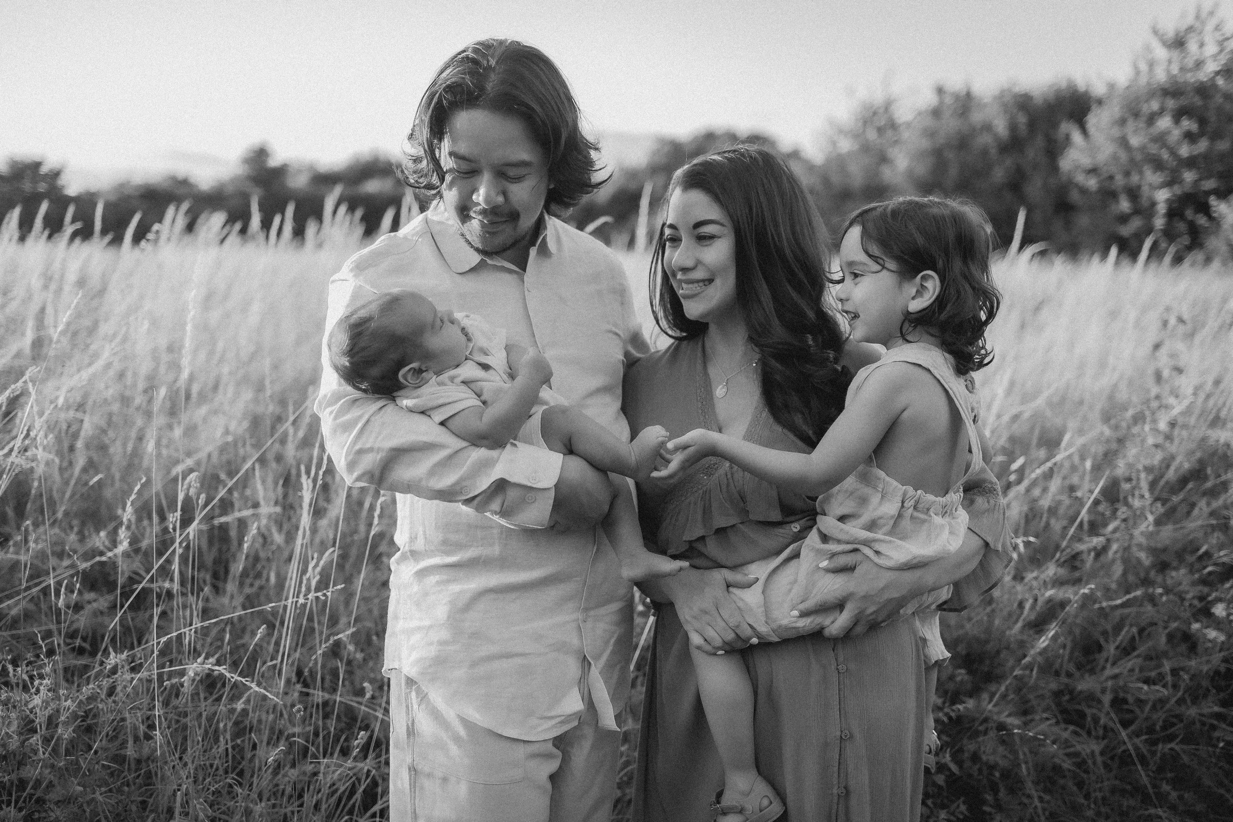 A family of four standing in a field, with the father holding a newborn baby, the mother holding a young girl, and all of them smiling at each other.