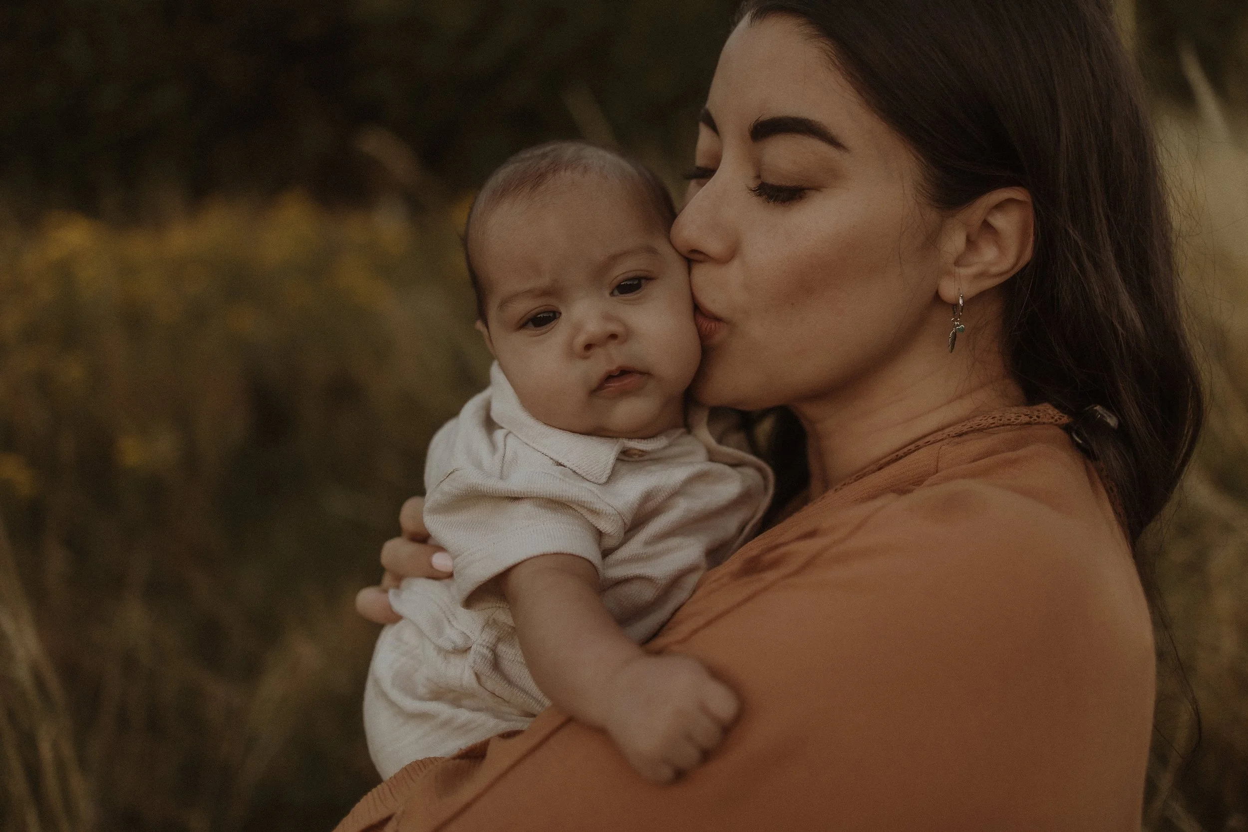 A woman holding a baby close, giving a gentle kiss on the baby's cheek in an outdoor setting with blurred natural background.