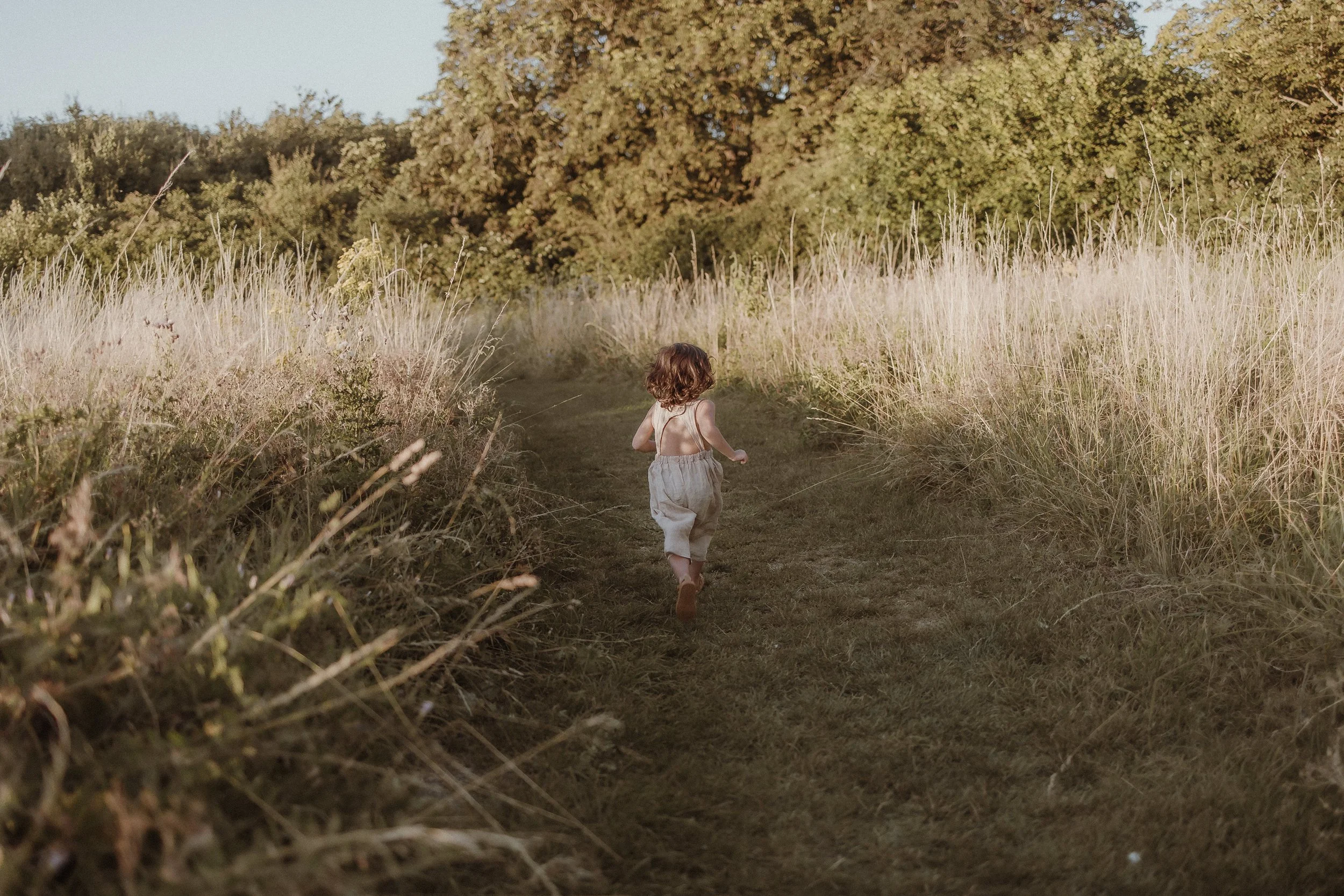 A young girl with brown hair, wearing beige overalls and a sleeveless top, running down a grassy dirt path surrounded by tall grasses and trees in the distance during late afternoon or early evening.