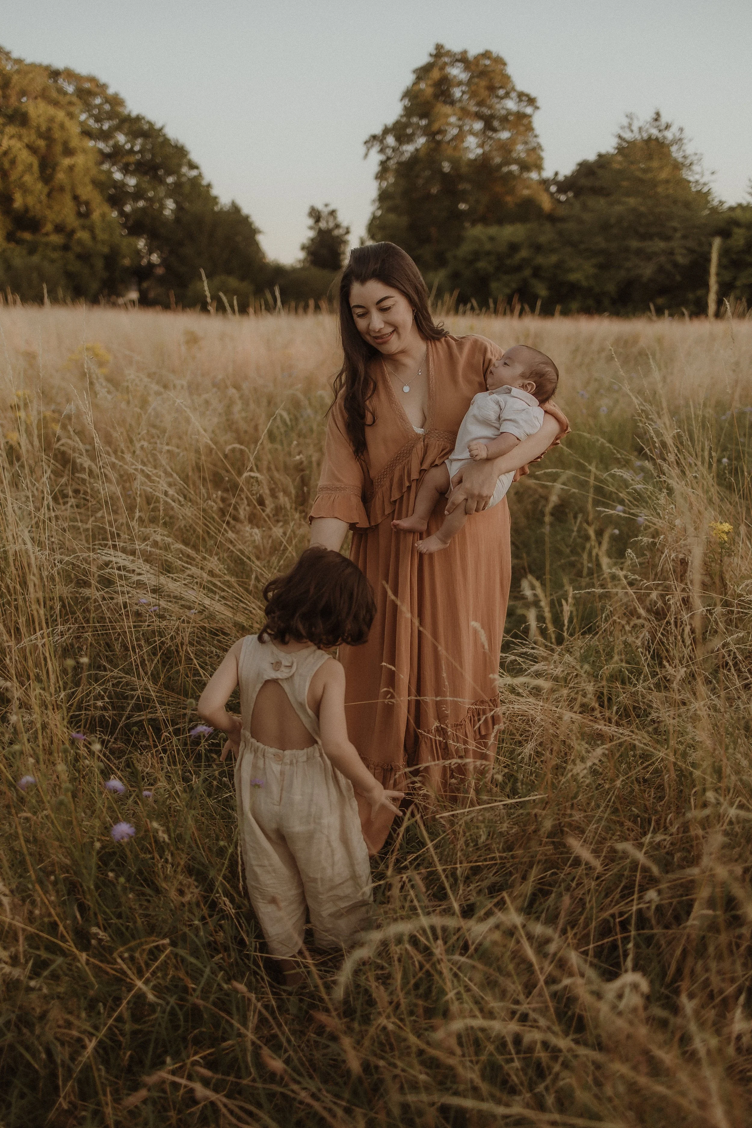 A woman holding a baby in a field of tall grass and wildflowers with two young children nearby, during sunset, with trees in the background.